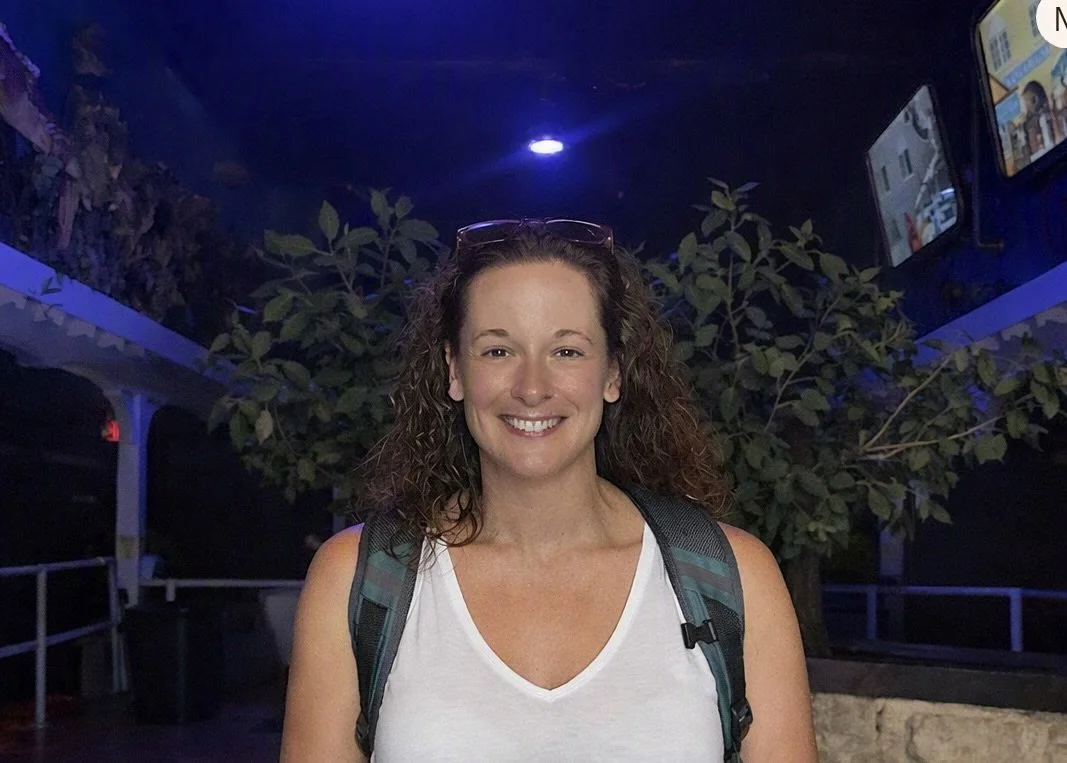 A woman with curly brown hair, wearing a white sleeveless top and glasses on her head, standing outdoors at night with a backpack, smiling at the camera, with greenery and lit screens in the background.