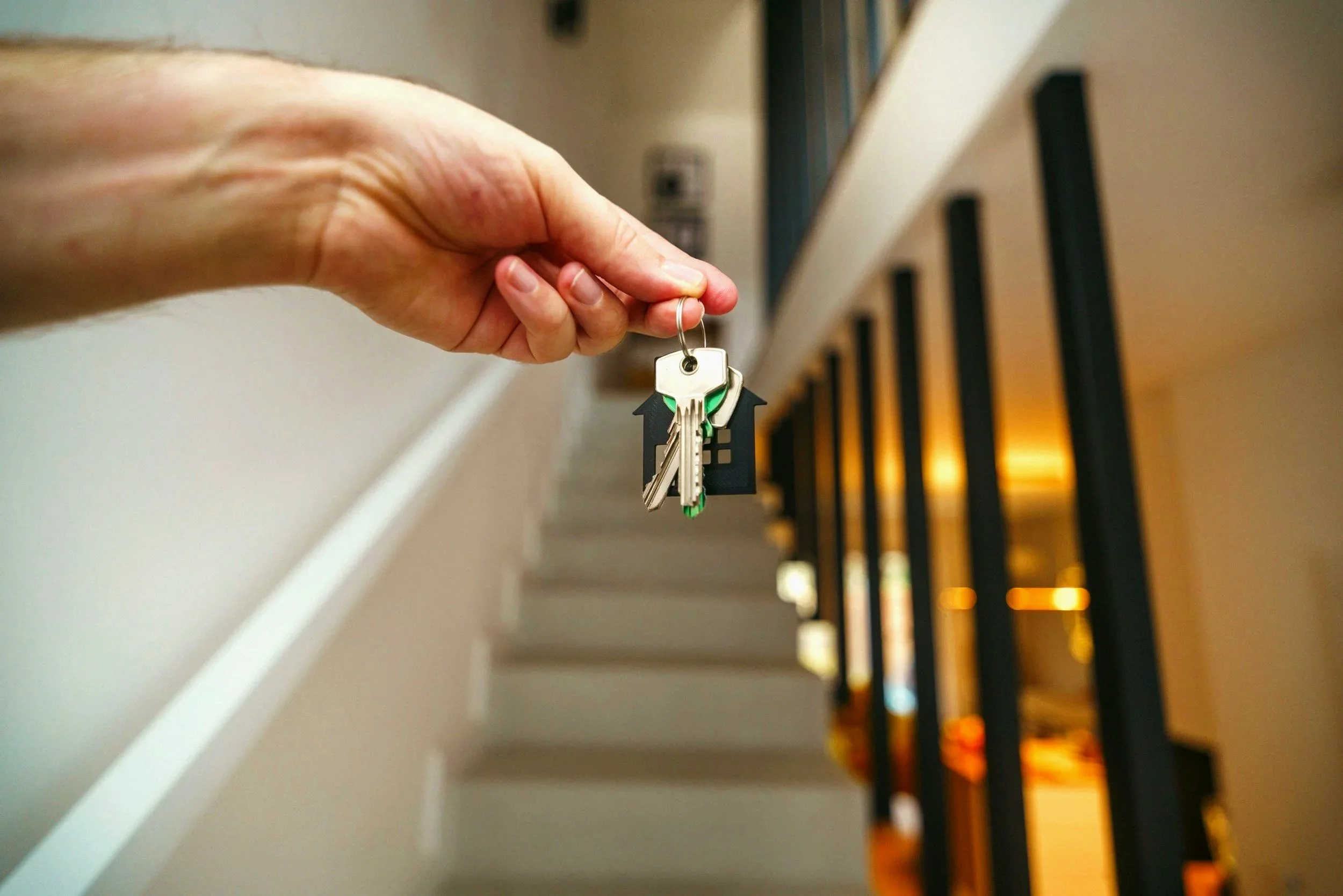 A hand holding keys with house-shaped keychains against a blurred background of a staircase and warm lighting.