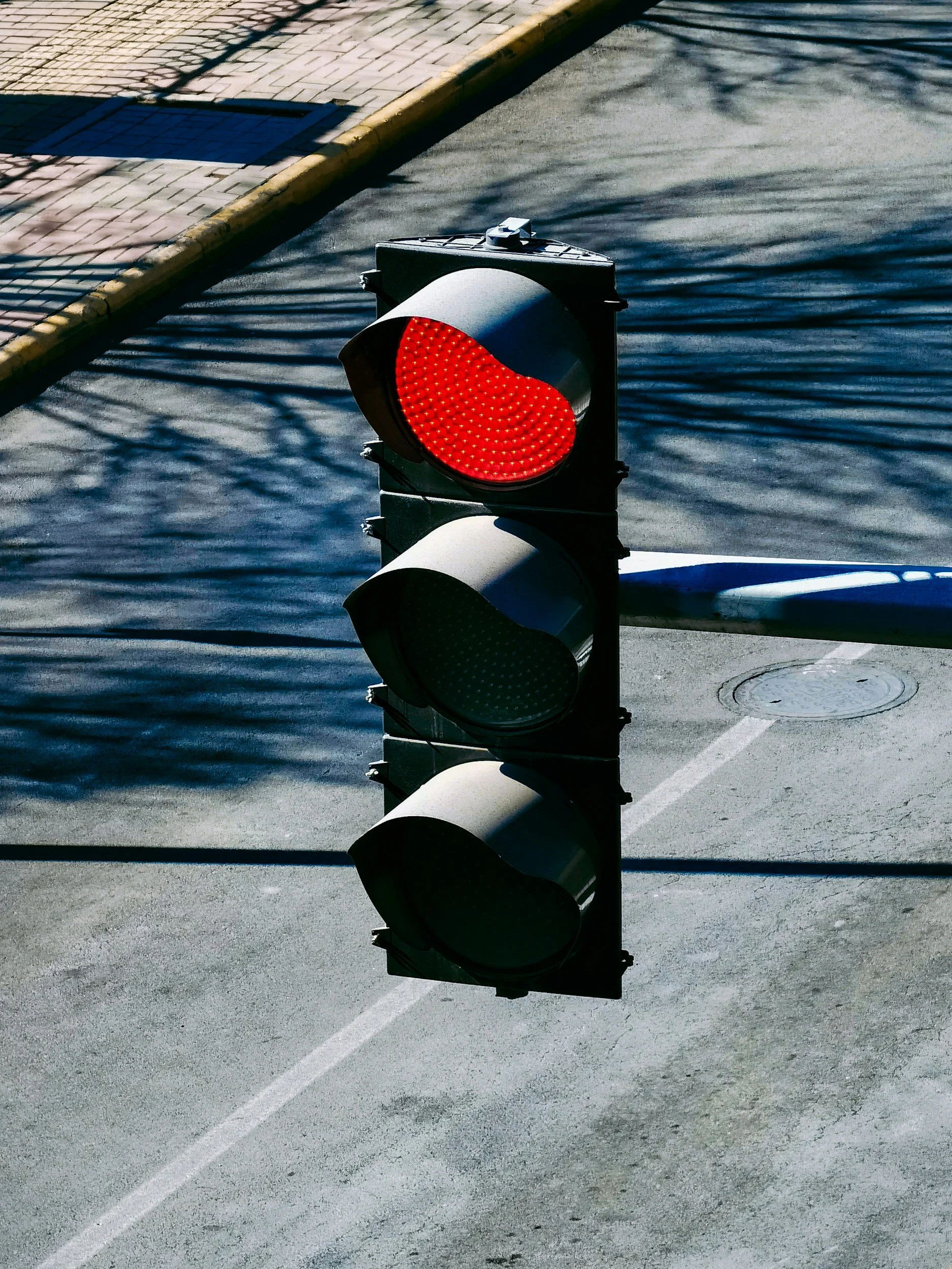 A traffic light displaying a red signal, situated on an urban street with shadow patterns and a sidewalk in the background.