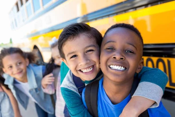 Smiling children hugging and walking near a school bus, with other kids in the background.