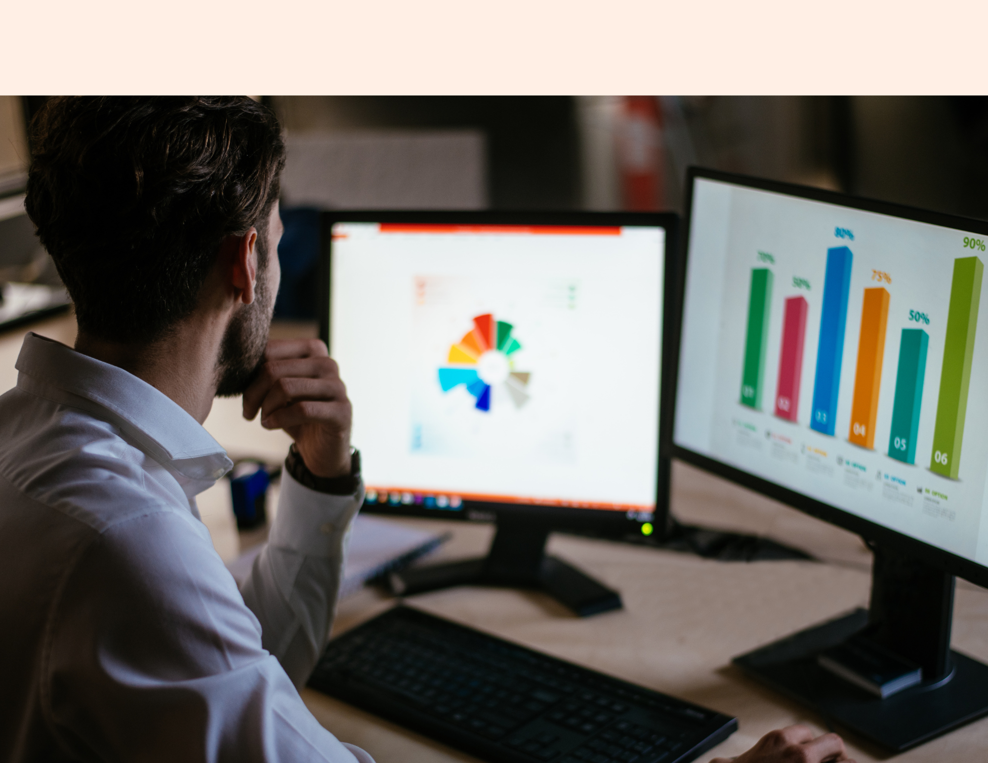 A man working at his desk with dual computer monitors displaying colorful bar charts and a pie chart.