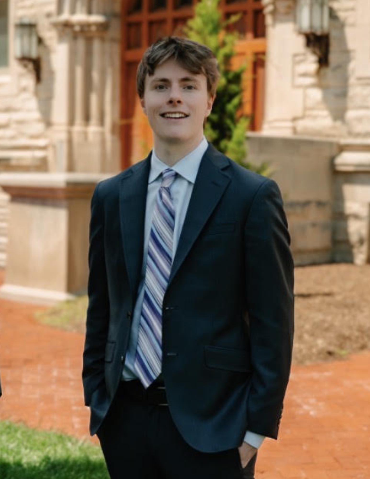 A young man dressed in a navy suit with a striped tie, standing outdoors in front of a historic building with stone columns and greenery, smiling at the camera.