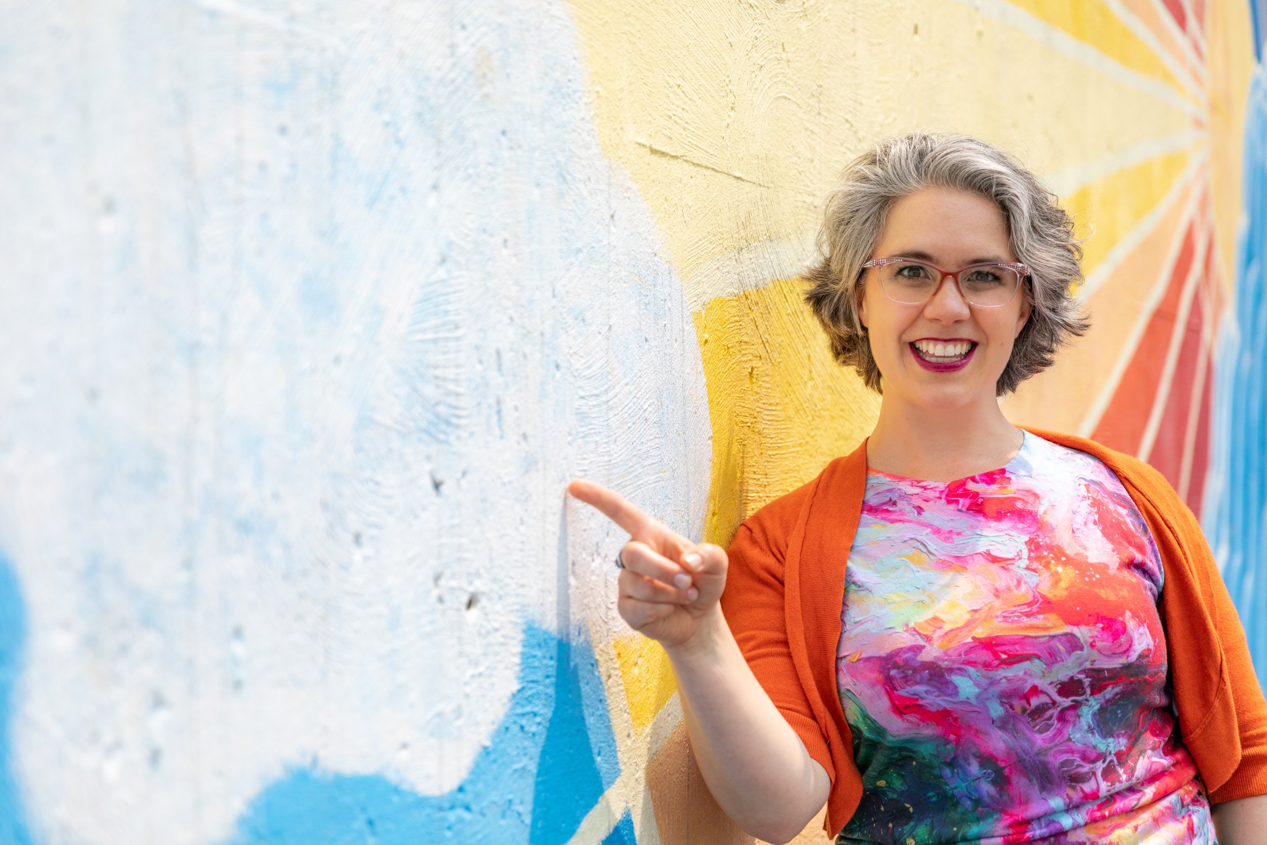 A woman with short gray hair, glasses, and a bright smile standing against a colorful mural wall, pointing to her left.