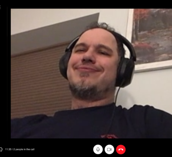 Man wearing headphones, smiling in a video call, with a framed picture on the wall behind him.