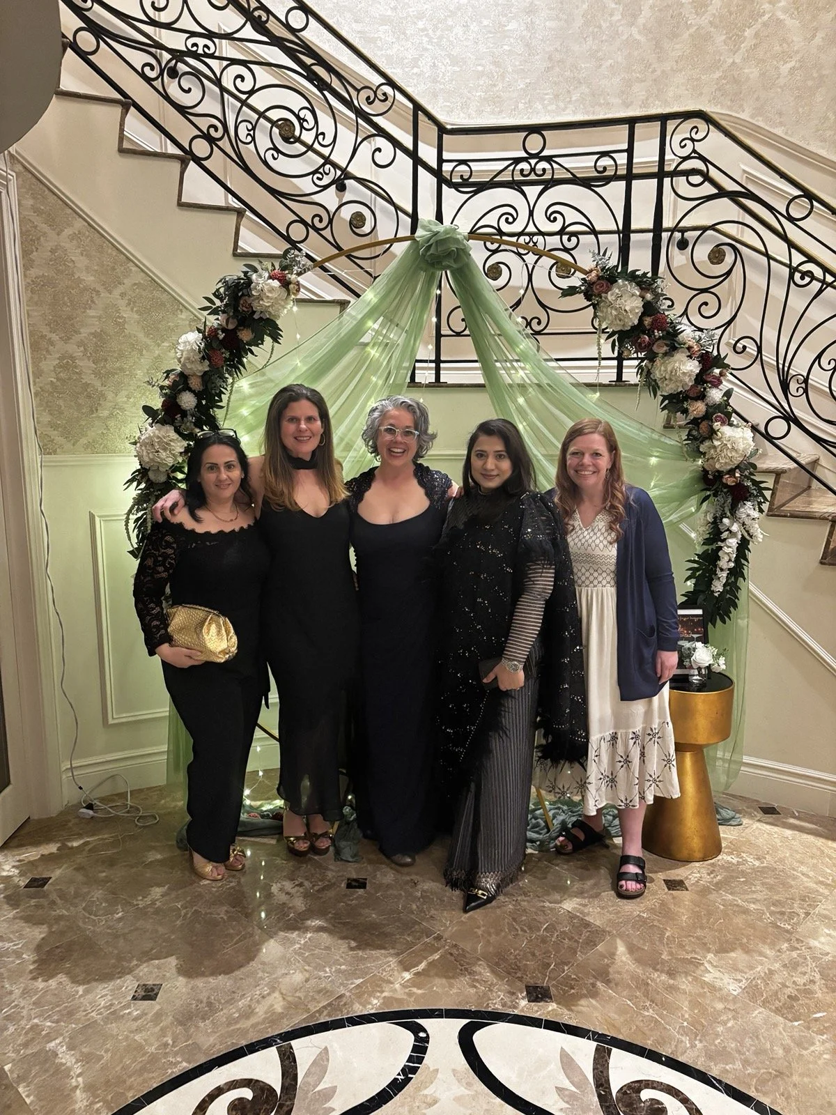 Five women standing together in front of a decorative floral arch with green drapery, at an indoor event, possibly a wedding or celebration, on a marble floor with a black and white patterned border.