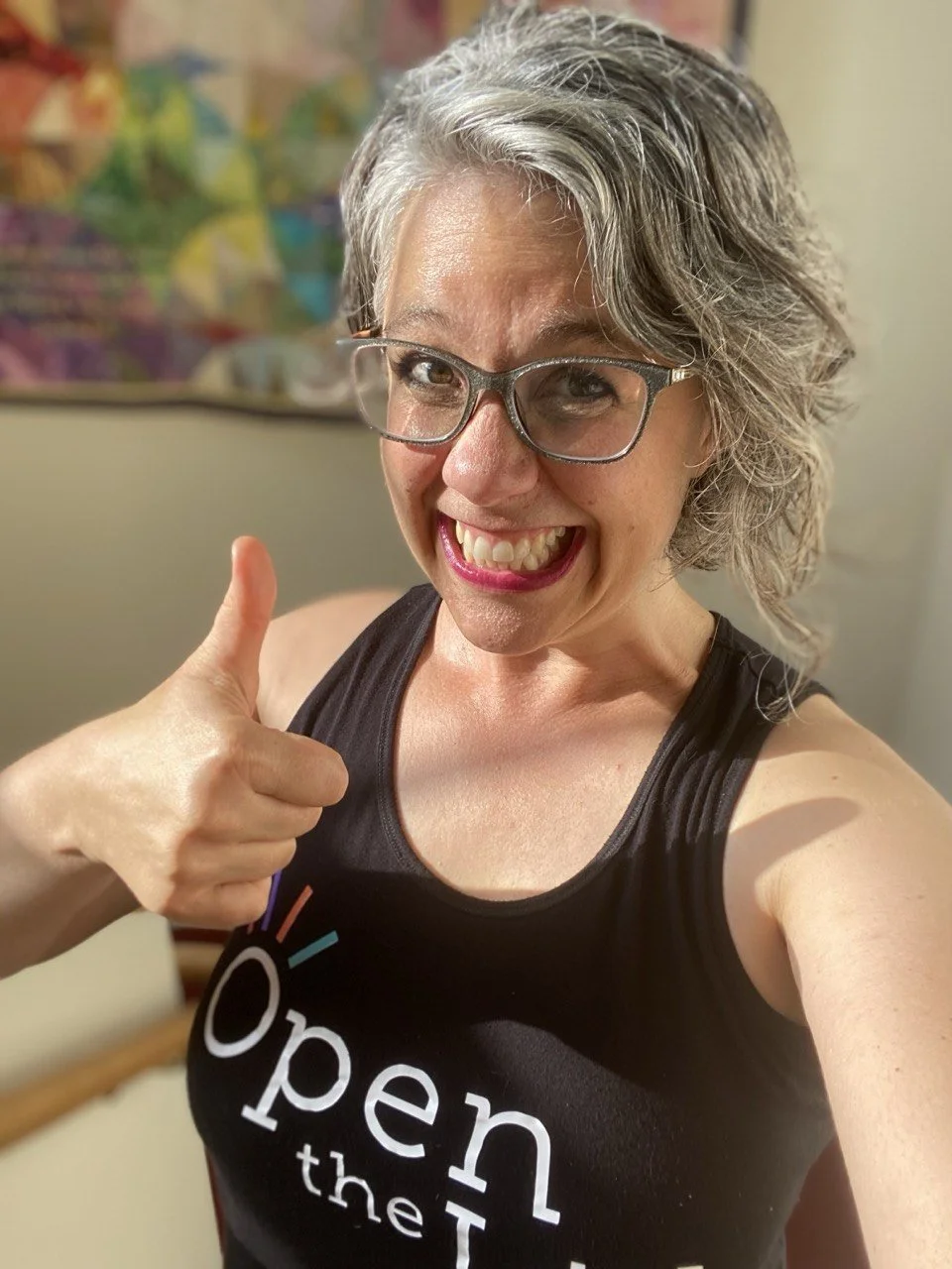 Smiling woman with gray hair and glasses giving a thumbs-up, wearing a black tank top that says 'Open the' with a colorful logo on it.