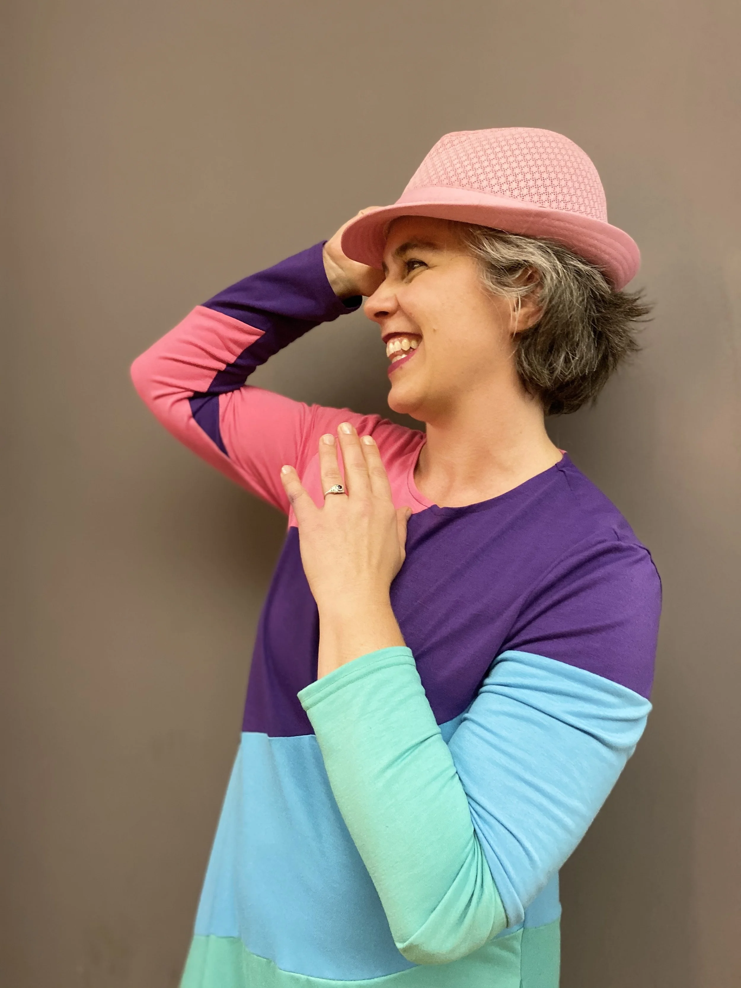 A woman with short grey hair smiling, wearing a pink hat with a textured pattern and a long-sleeved shirt with rainbow colors, standing against a neutral background.