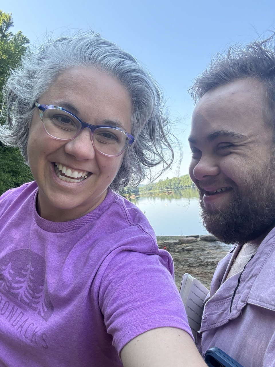 A woman with gray hair and glasses smiling for a selfie with a man with a beard, both outdoors near a lake with trees and a blue sky in the background.