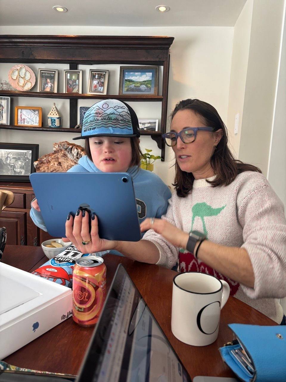 Woman and boy sitting at a table looking at a tablet. The woman is wearing glasses and a sweater, and the boy is wearing a cap and a blue hoodie. There are snacks, a can of sparkling water, and laptops on the table, with a bookshelf and framed photos in the background.