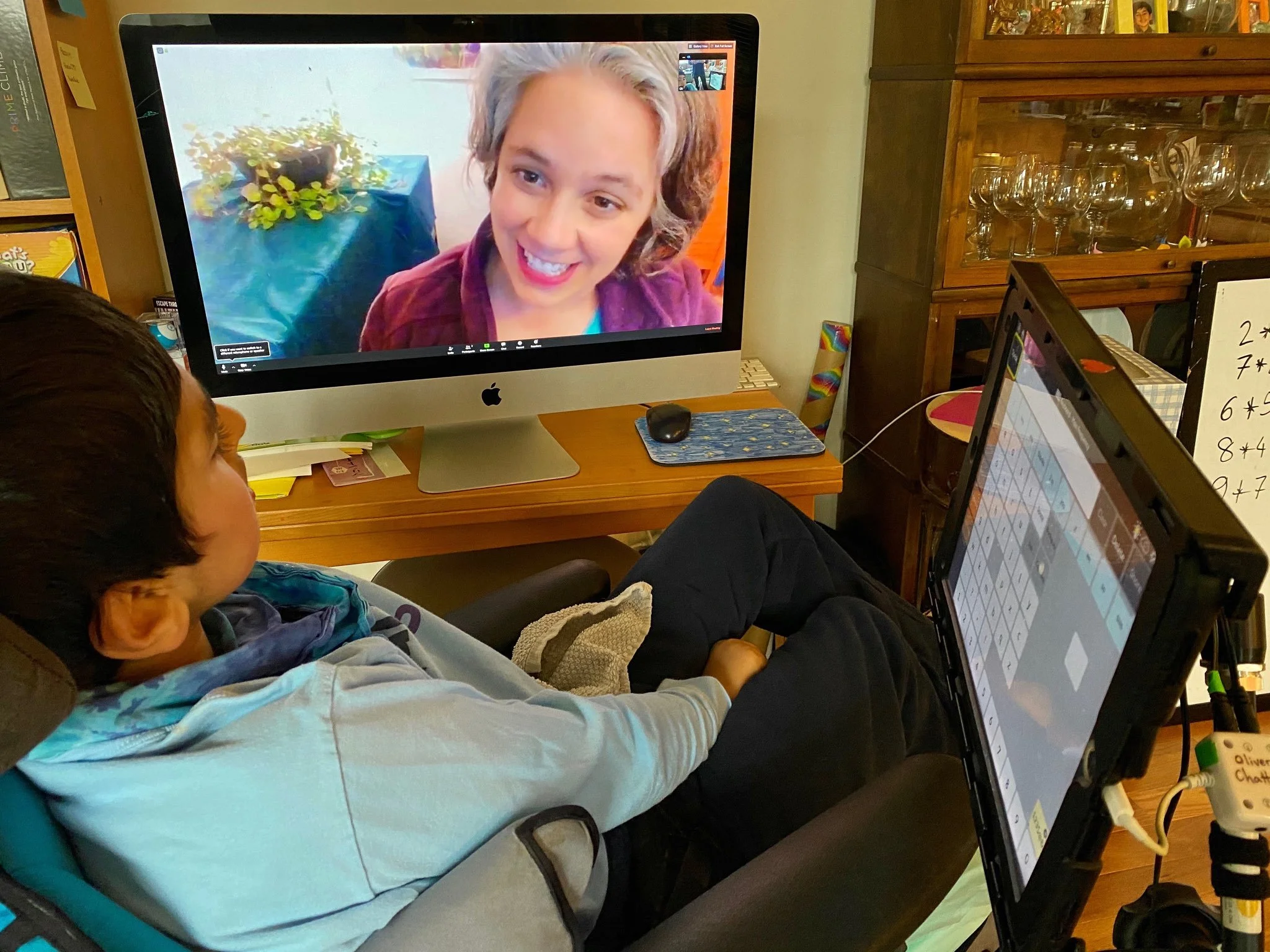 A person in a wheelchair participating in a video call with a woman with short gray hair, smiling. The scene is set indoors with wooden furniture, glasses, and a computer monitor showing the video call.