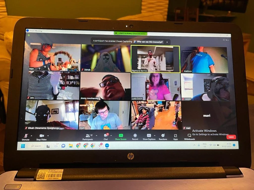 Zoom video conference with multiple participants, some in household settings, some with headphones, and interactive backgrounds. The screen shows icons for controls and a green notification indicating the computer is sharing. The laptop is in a room with a green wall and wooden floor.