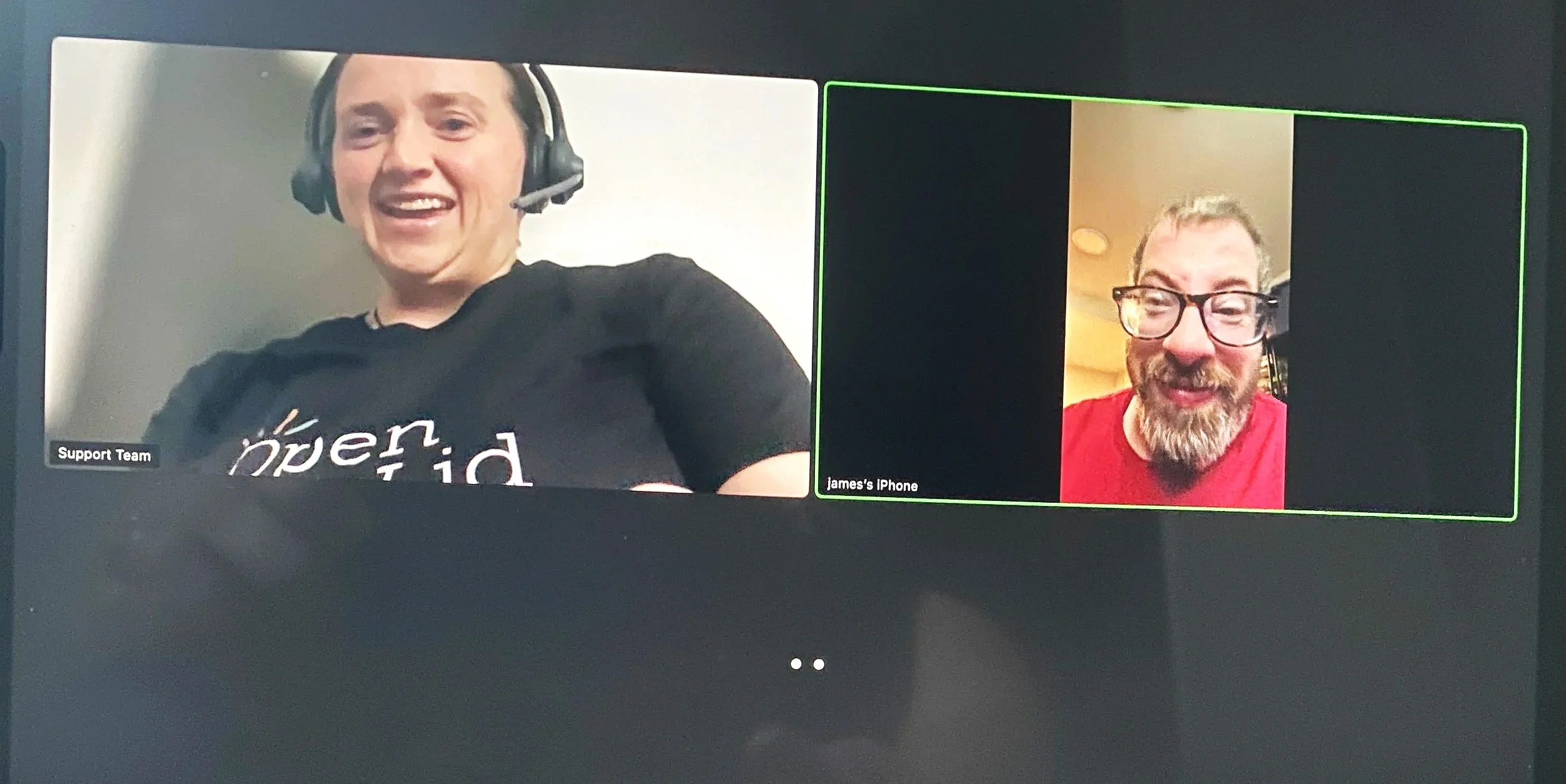 Two men are seen on a video call. One on the left is wearing a black shirt, headphones, and a headset microphone, smiling. The other on the right wears glasses and a red shirt, also smiling. The screen displays labels "Support Team" and "James's iPhone."