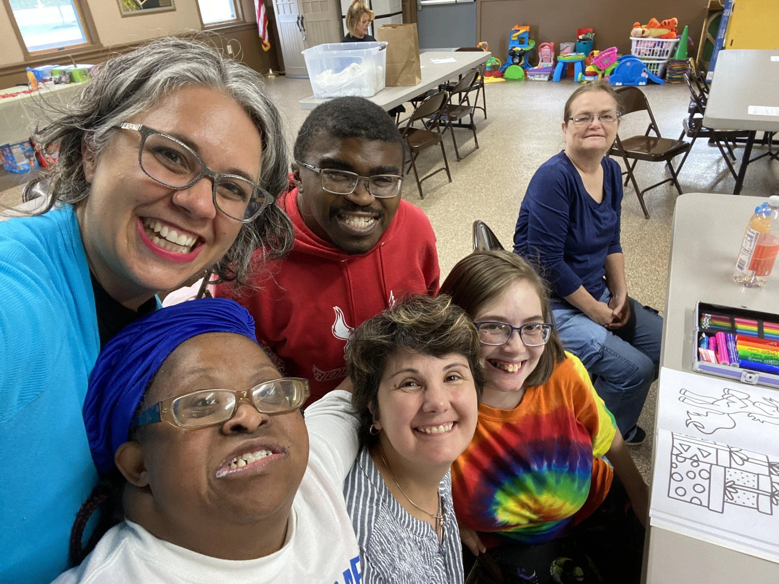 A group of smiling people taking a selfie in a room with tables and children's toys in the background. The group is diverse in age and ethnicity, and they are wearing colorful casual clothing.