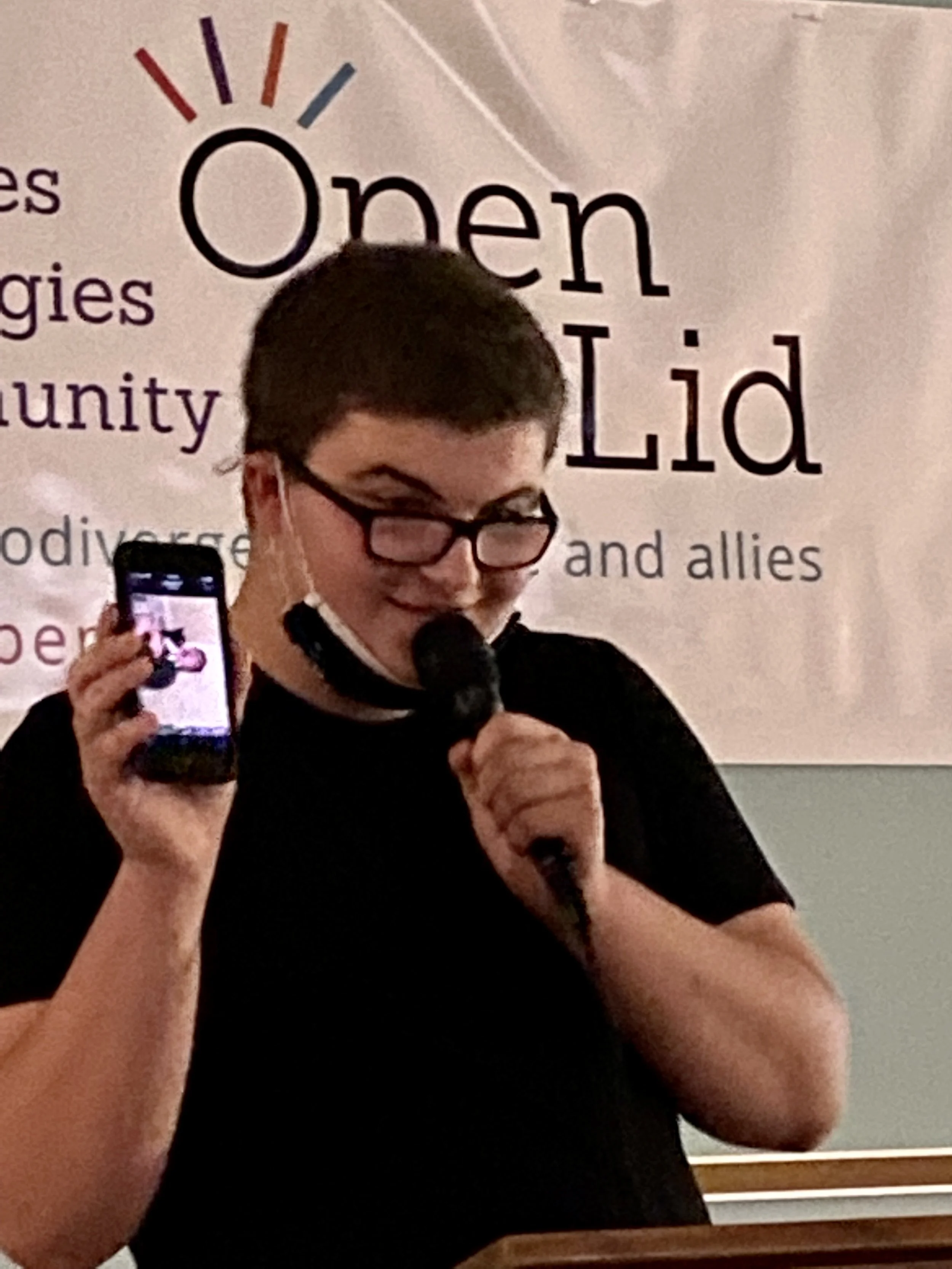 Person with short dark hair and glasses speaking into a microphone while holding a smartphone, in front of a backdrop with text that reads "Open" and other words related to community and allies.