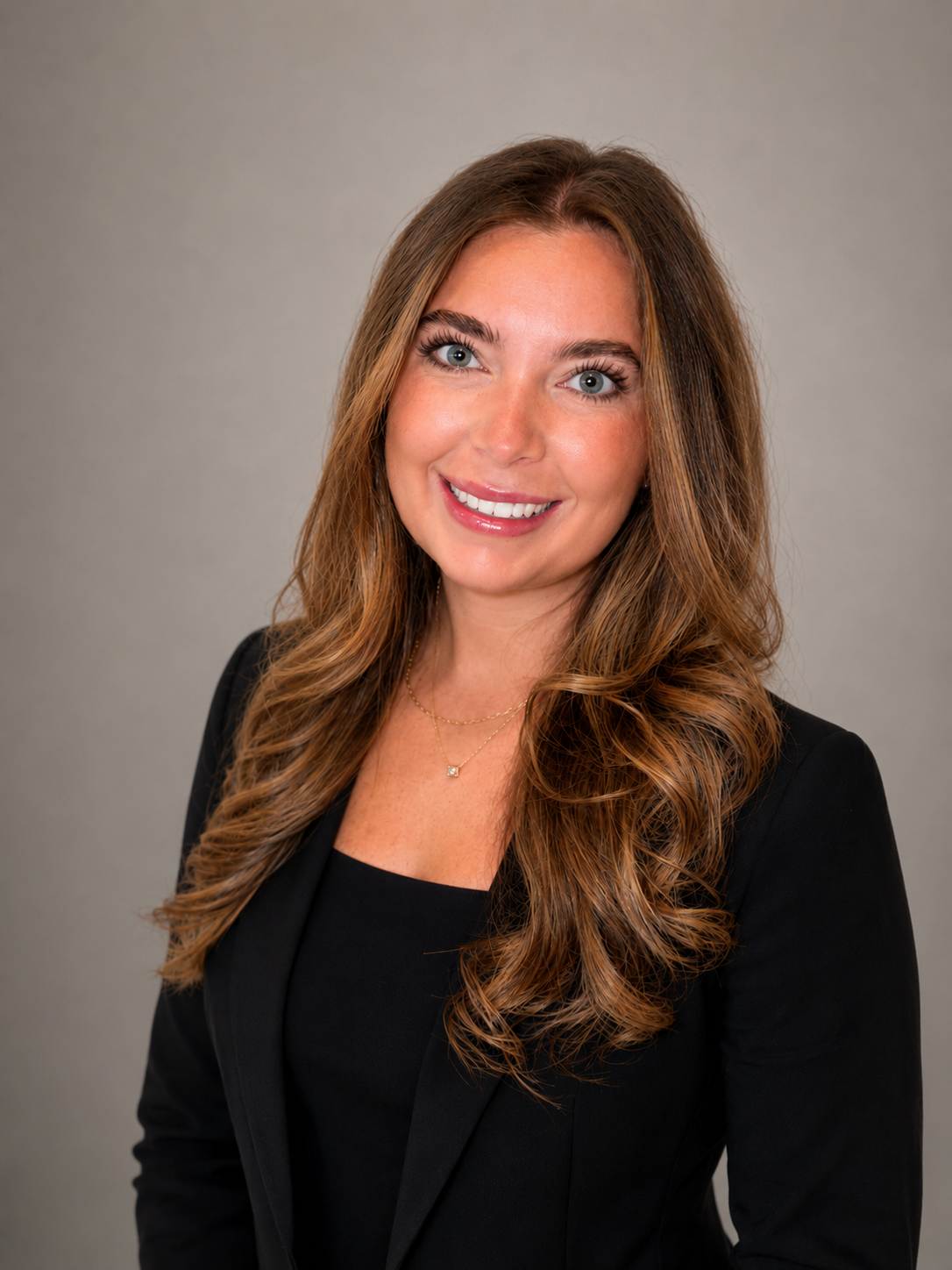 A woman with long, curly, light brown hair wearing a black blazer and a gold necklace, smiling against a gray background.
