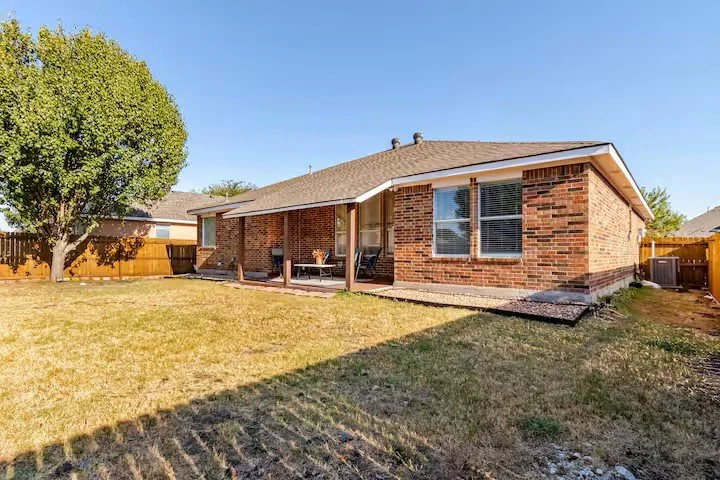 Backyard of a single‑story brick house with a covered patio, wooden fence, tree on the left, and clear blue sky.