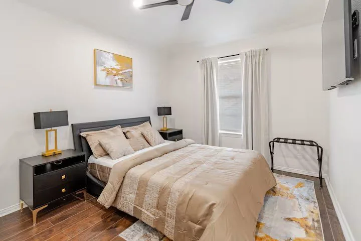 Modern bedroom with bed, black nightstands, yellow lamps, gold and white abstract painting, white curtains, rug, wall‑mounted TV, and ceiling fan.