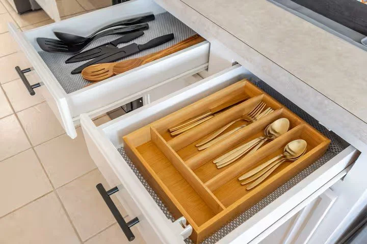 Open kitchen drawers showing black plastic utensils in one drawer and bamboo utensils with matching cups in another.