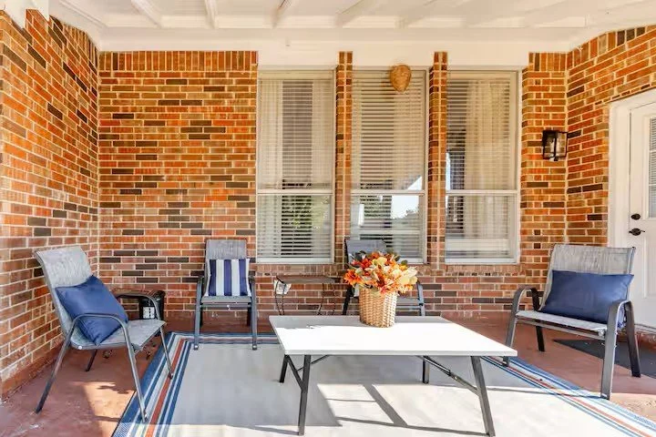 Back porch with brick walls, three windows with white blinds, outdoor furniture including a white table with flowers, blue‑cushioned chairs, wooden chair with striped cushion, and wall light fixture.