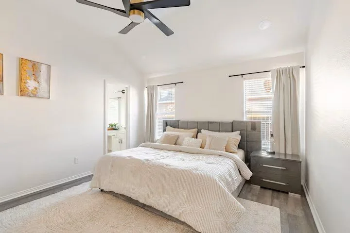 Bright bedroom with a large bed, gray upholstered headboard, white bedding, multiple pillows, two windows with white curtains, gray nightstands, and wall art.