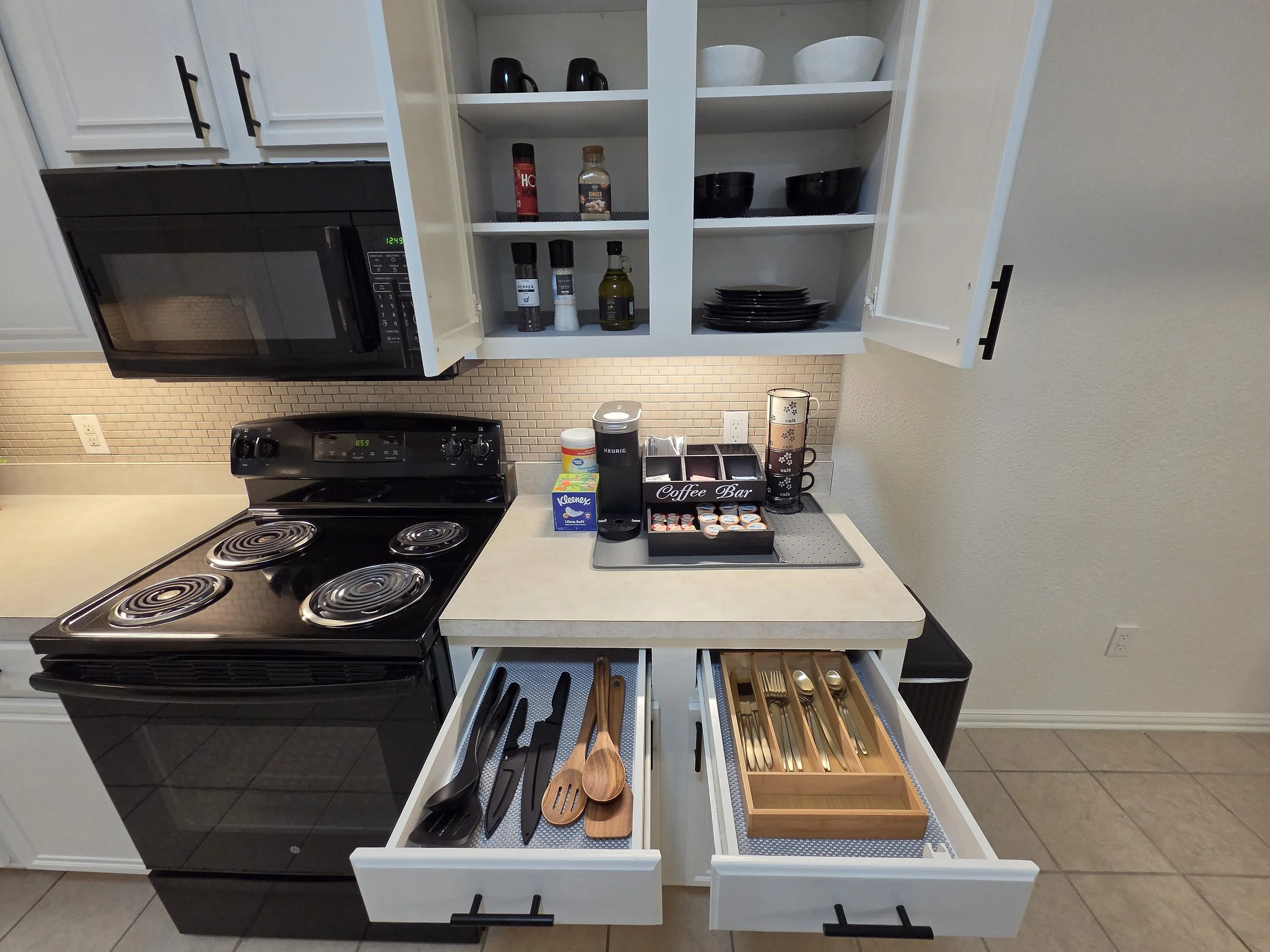 A kitchen drawer opened to reveal wooden and black plastic utensils, with a pull-out spice rack containing small containers, and a section with silverware in a tray.