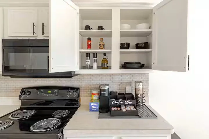 Kitchen coffee bar with open white cabinets, black stove, coffee capsules, mugs, and accessories.