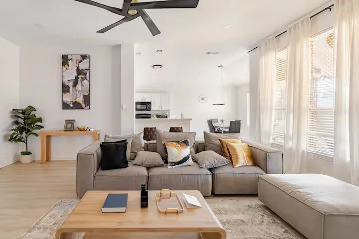 Living room with a gray sectional sofa, colorful pillows, wooden coffee table, sheer curtains, ceiling fan, and an open kitchen and dining area.