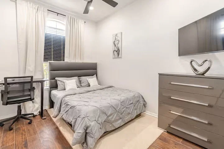 Bedroom with gray quilted bed, four pillows, gray headboard, window with white curtains, black desk and chair, gray dresser with metallic handles, wall‑mounted TV, decorative sculpture, and wall art.
