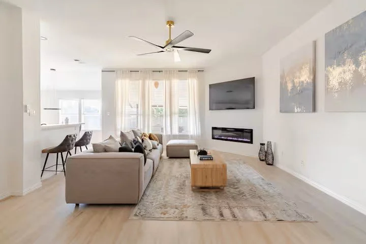 Modern living room with gray sofa, wooden coffee table, flat‑screen TV above an electric fireplace, abstract wall art, and decorative vases.