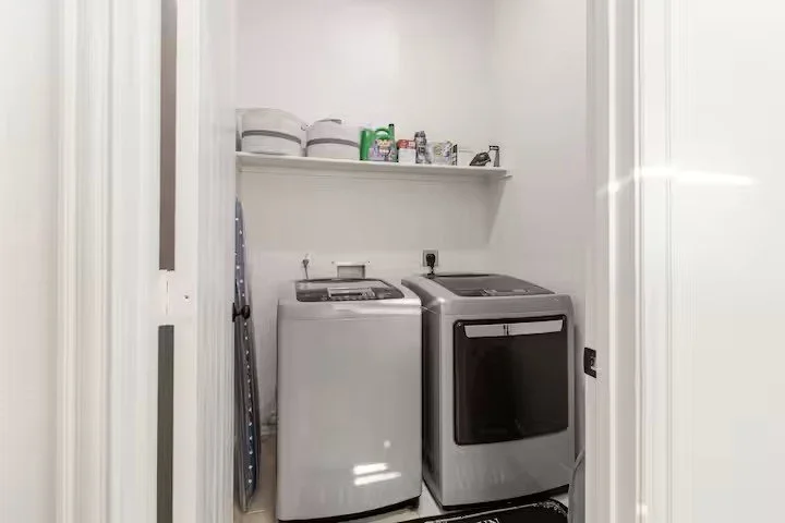 Laundry room with top‑loading washing machine, front‑loading dryer, shelf holding laundry supplies, and ironing board against the wall.