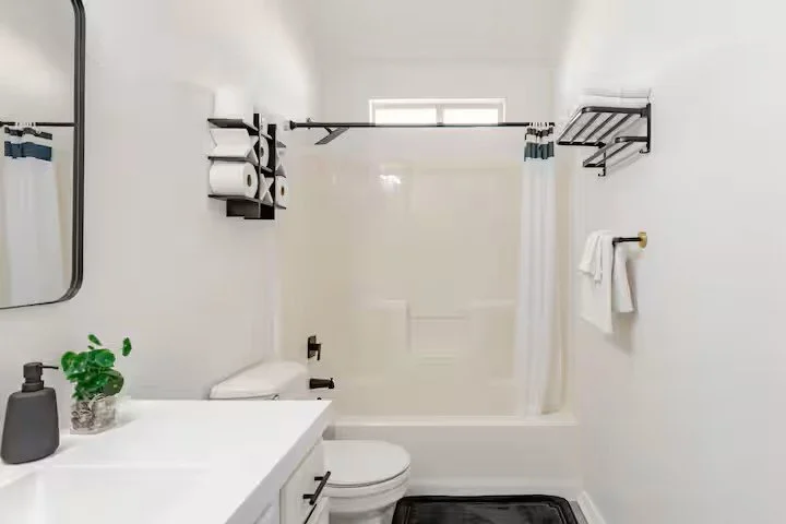 Clean white bathroom with vanity, mirror, toilet, bathtub with shower curtain, wall‑mounted shelves holding towels and toiletries, and a small high window.