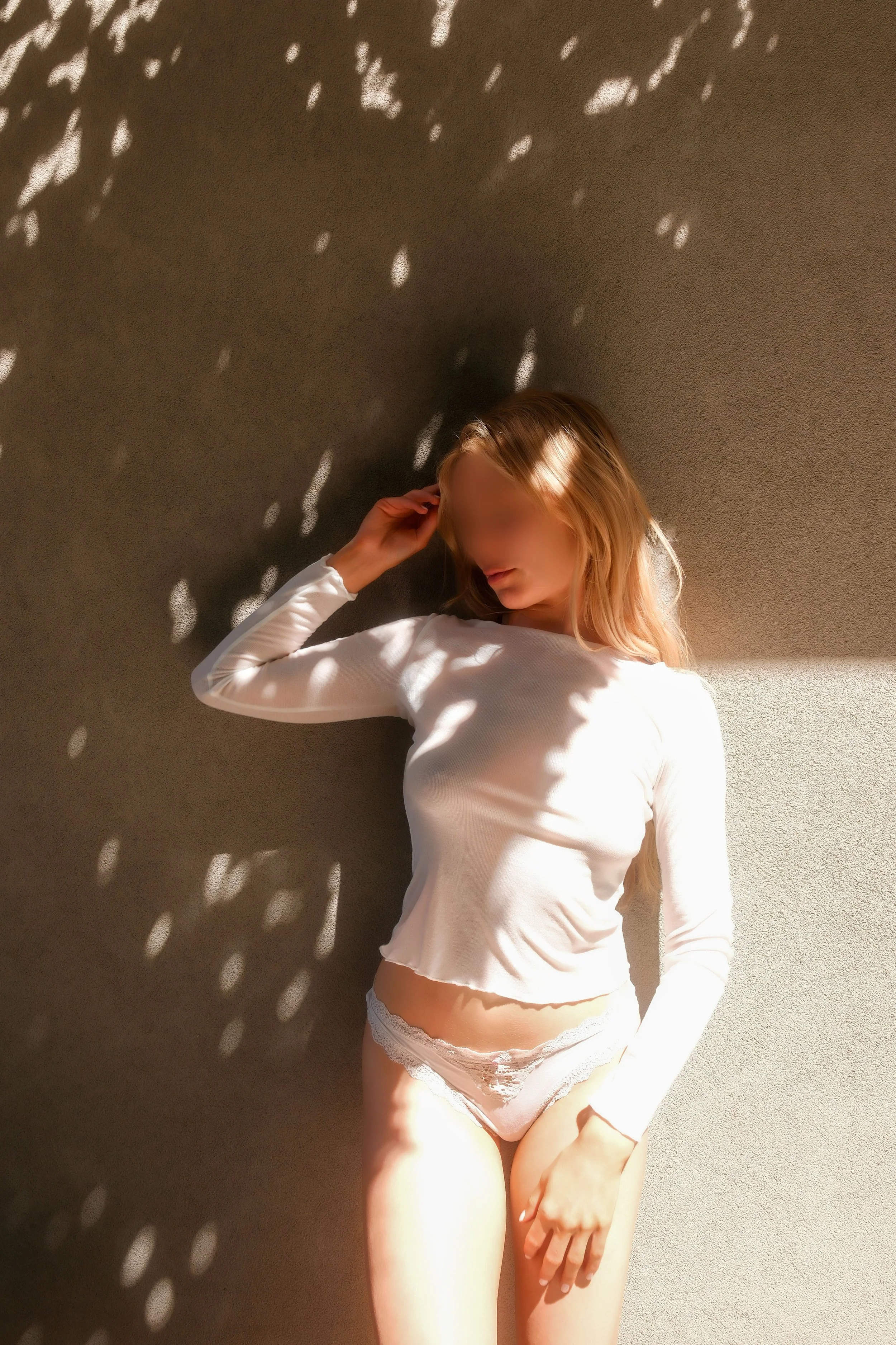 A woman with long blonde hair lying on her side on a surface, wearing a white long-sleeve shirt and lace underwear, with sunlight creating shadow patterns on the wall behind her.