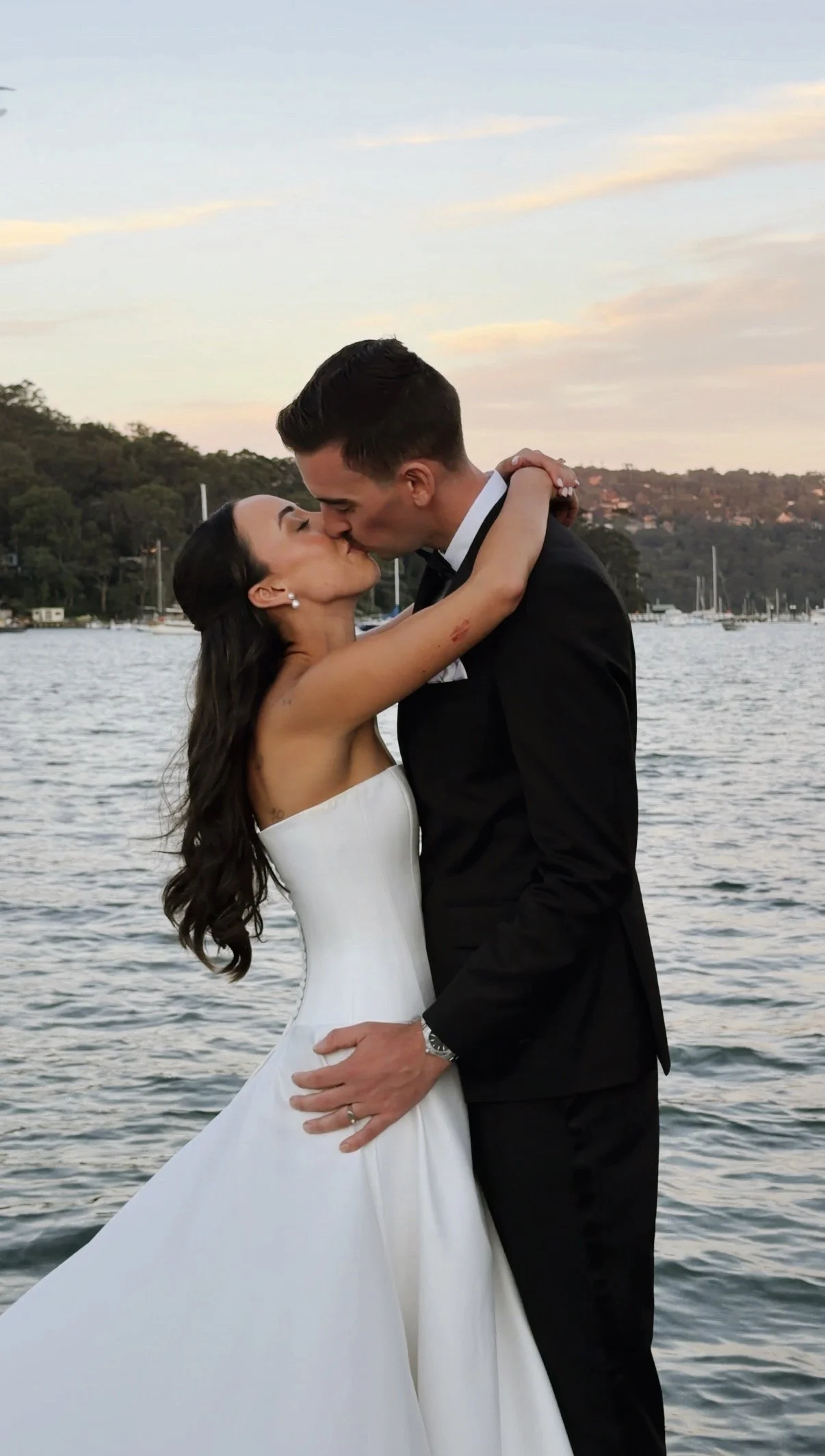 A bride and groom kiss by a body of water during sunset, with sailboats and a hilly landscape in the background.