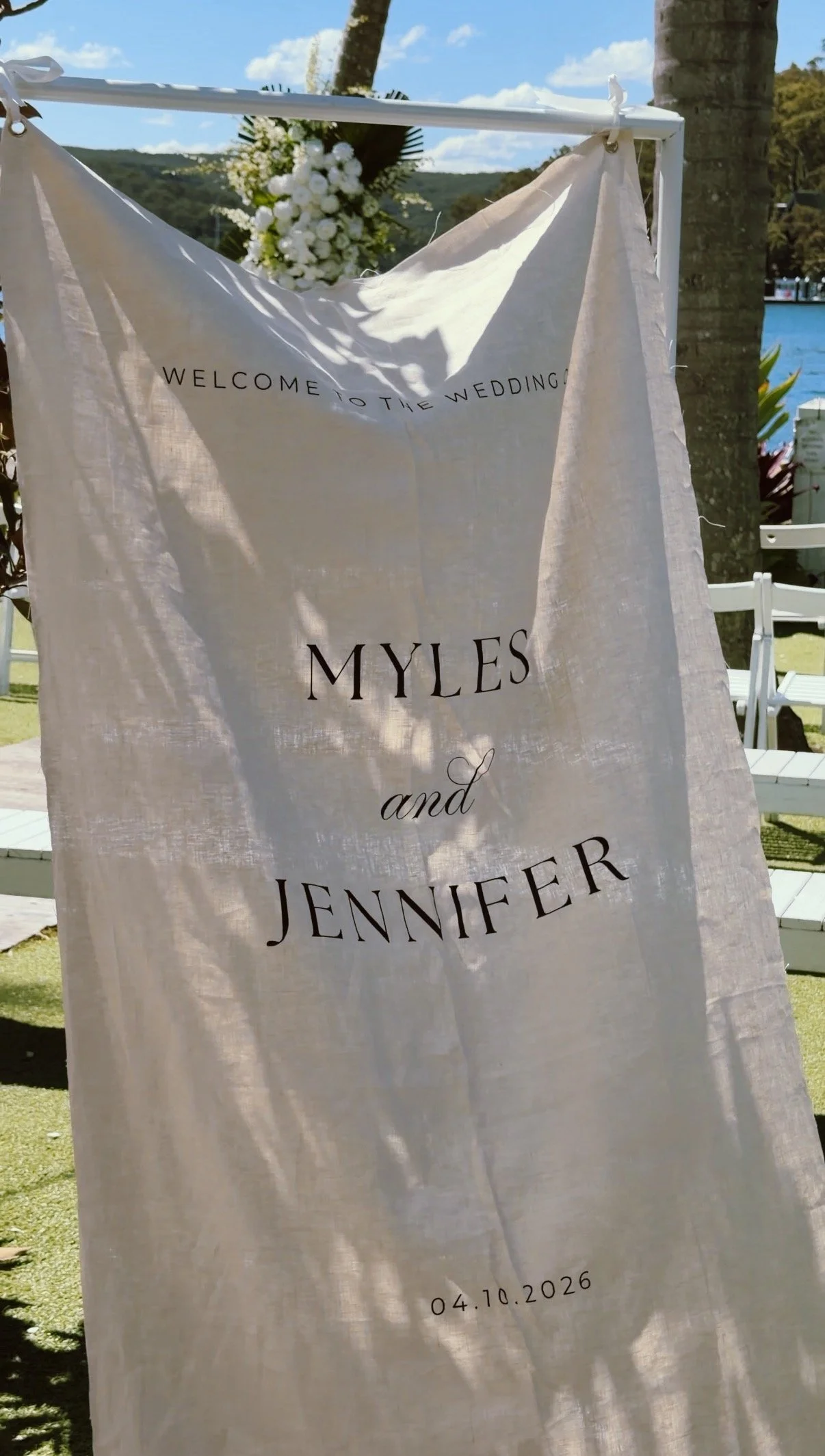 A wedding welcome banner made of light fabric with text welcoming guests to the wedding of Myles and Jennifer, scheduled for April 10, 2026, displayed outdoors with trees, flowers, and water in the background.