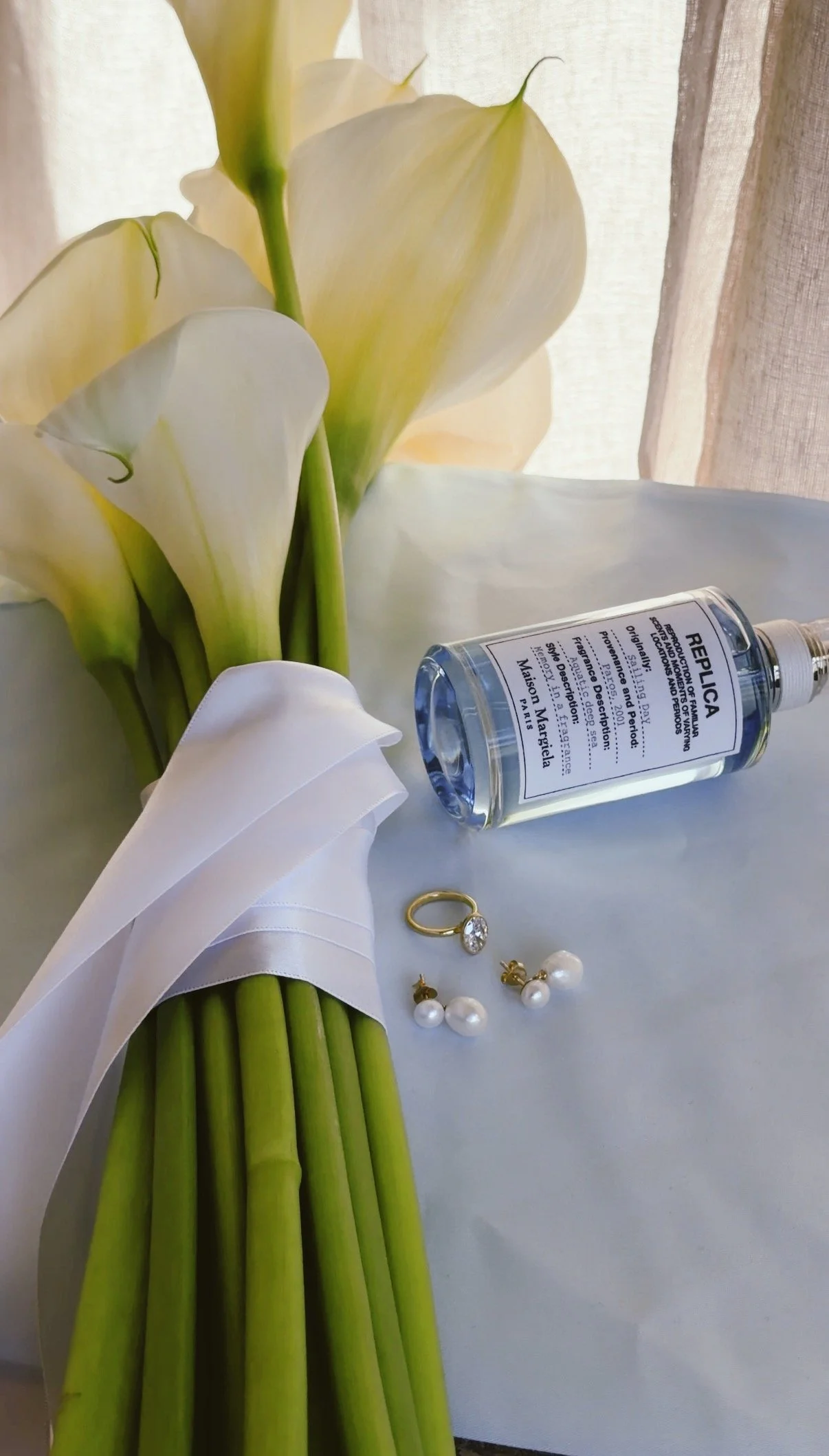 A bouquet of white calla lilies with a white ribbon wrapped around the stems, a pair of pearl earrings, a gold ring with a diamond, and a small bottle of Replika facial serum on a light blue surface, with a sheer curtain in the background.