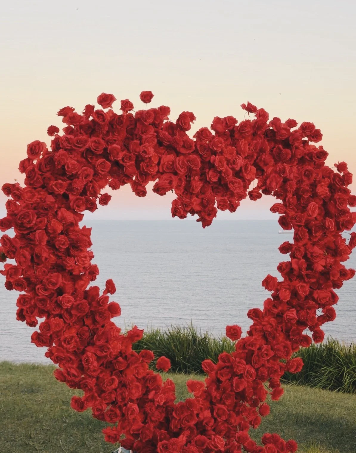 A large heart-shaped arch made of red roses, with a scenic ocean and sky background.