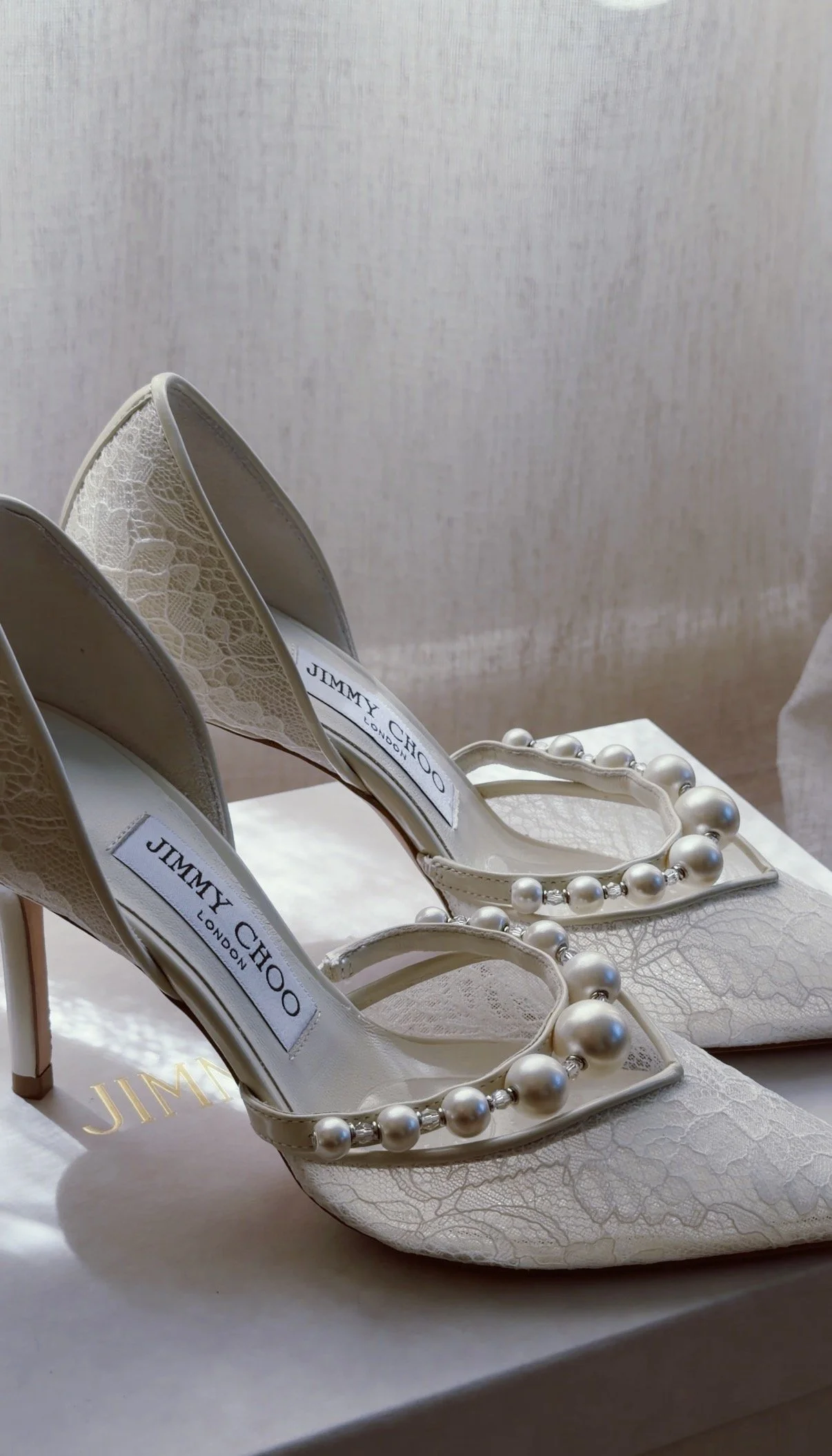 A pair of white Jimmy Choo high-heeled shoes with lace details and a pearl necklace decoration on the front, placed on a white surface.