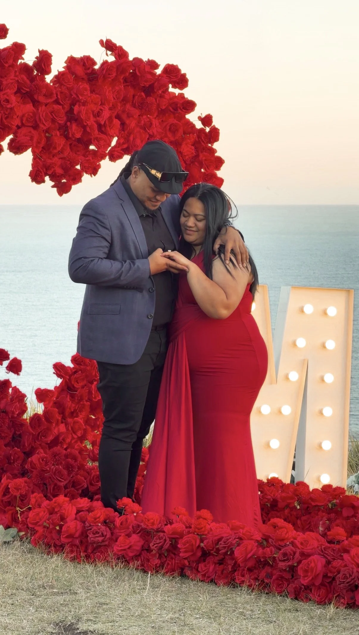 A couple stands together on a romantic outdoor setting by the water, surrounded by red roses and a large illuminated letter 'V'. The woman wears a red dress and the man wears a blue blazer and black pants, as they look at their phones and smile.