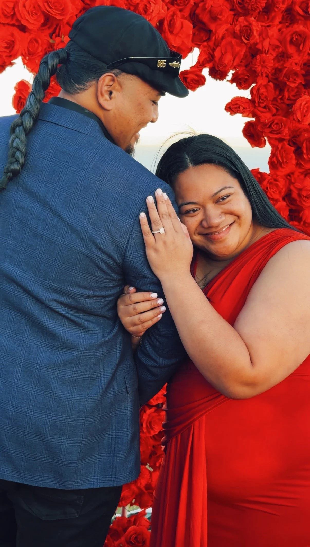 A couple embracing, with a backdrop of red roses. The woman is dressed in a red gown and is smiling, showing her engagement ring. The man is wearing a blue suit and a black cap.