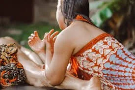 Woman in traditional clothing performing a Thai massage on a person lying face down outdoors