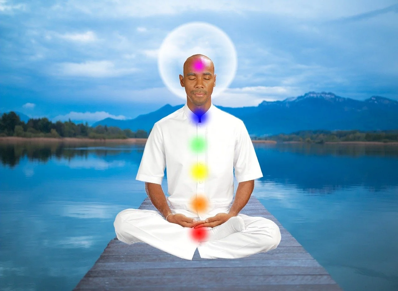 A man in meditation seated on a wooden dock by a lake, with a mountain landscape in the background. There are colored energy points along his body and a halo-like circle behind his head.
