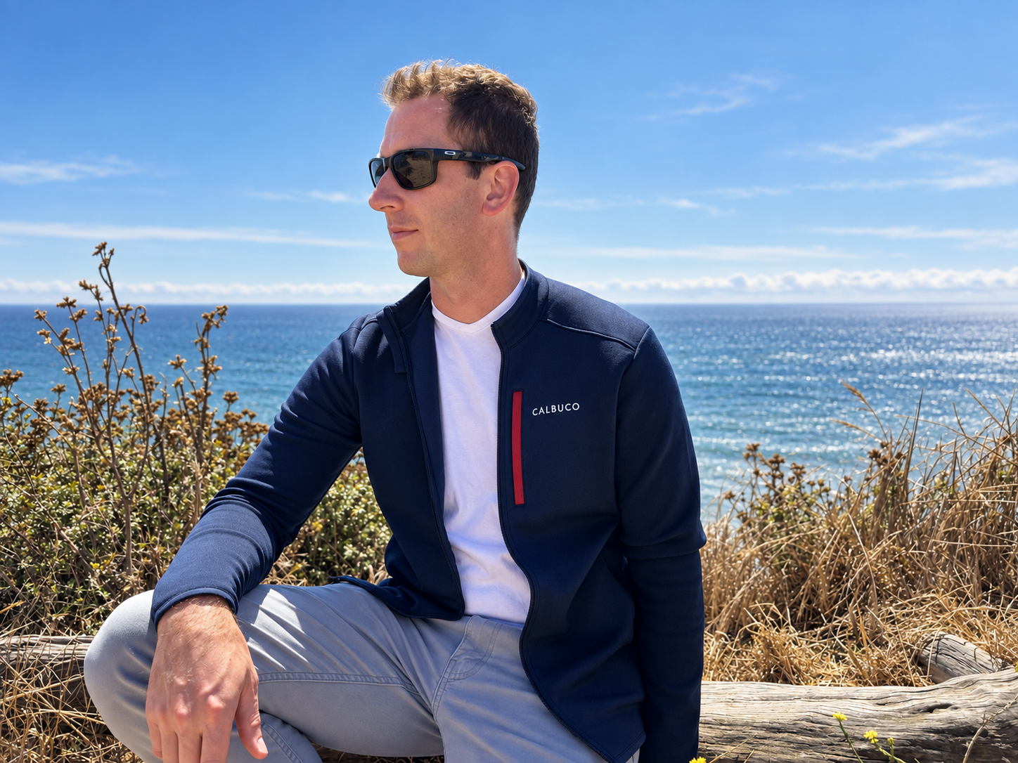 A man sitting outdoors near the ocean with dry bushes and grass around him, wearing sunglasses, a navy blue Calbuco jacket, a white t-shirt, and light gray pants, looking to the side with a blue sky and ocean in the background.