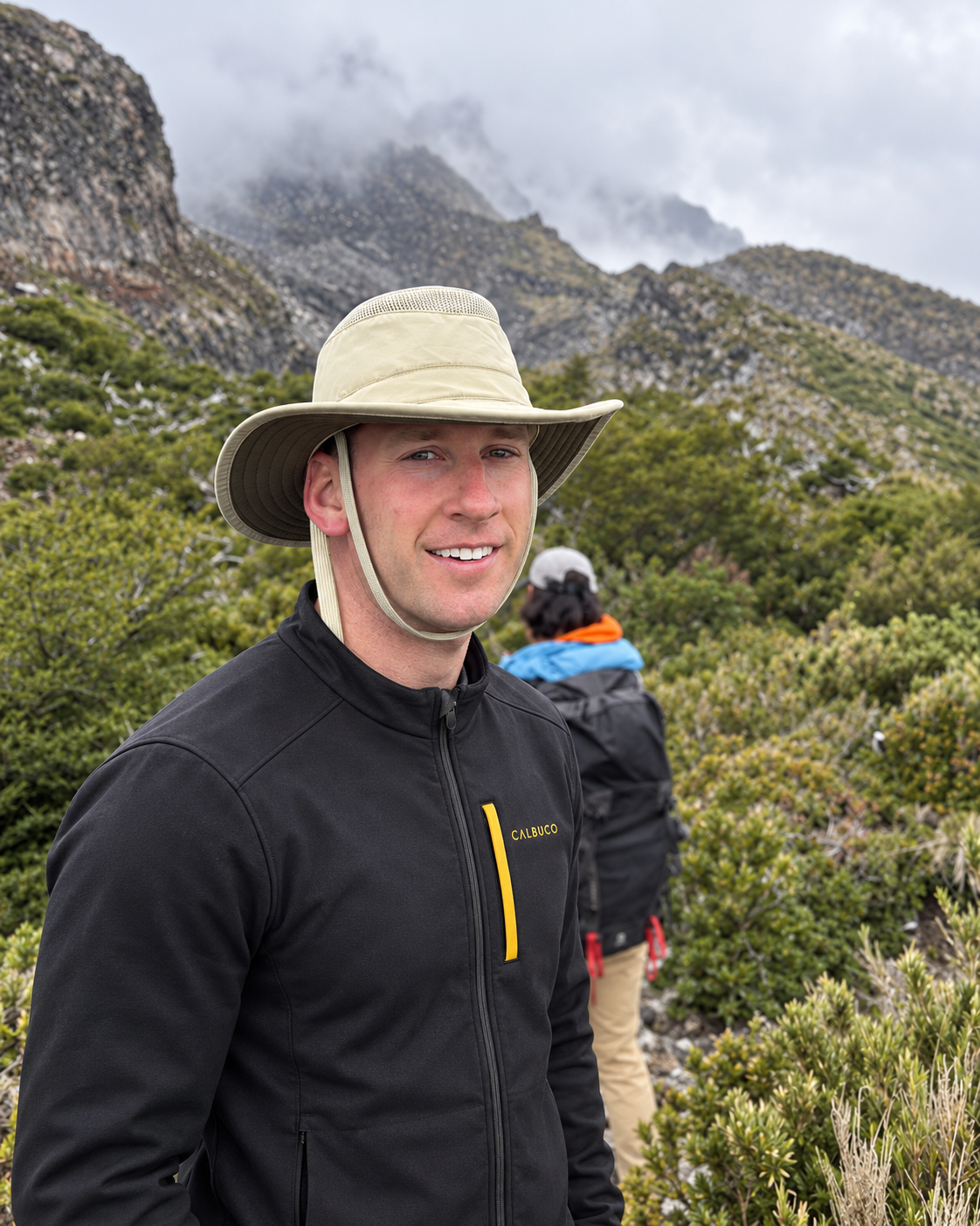A smiling man wearing a wide-brimmed hat and a black jacket with yellow accents stands outdoors in a mountainous, forested area with overcast skies. Another person with a backpack and cap is in the background.
