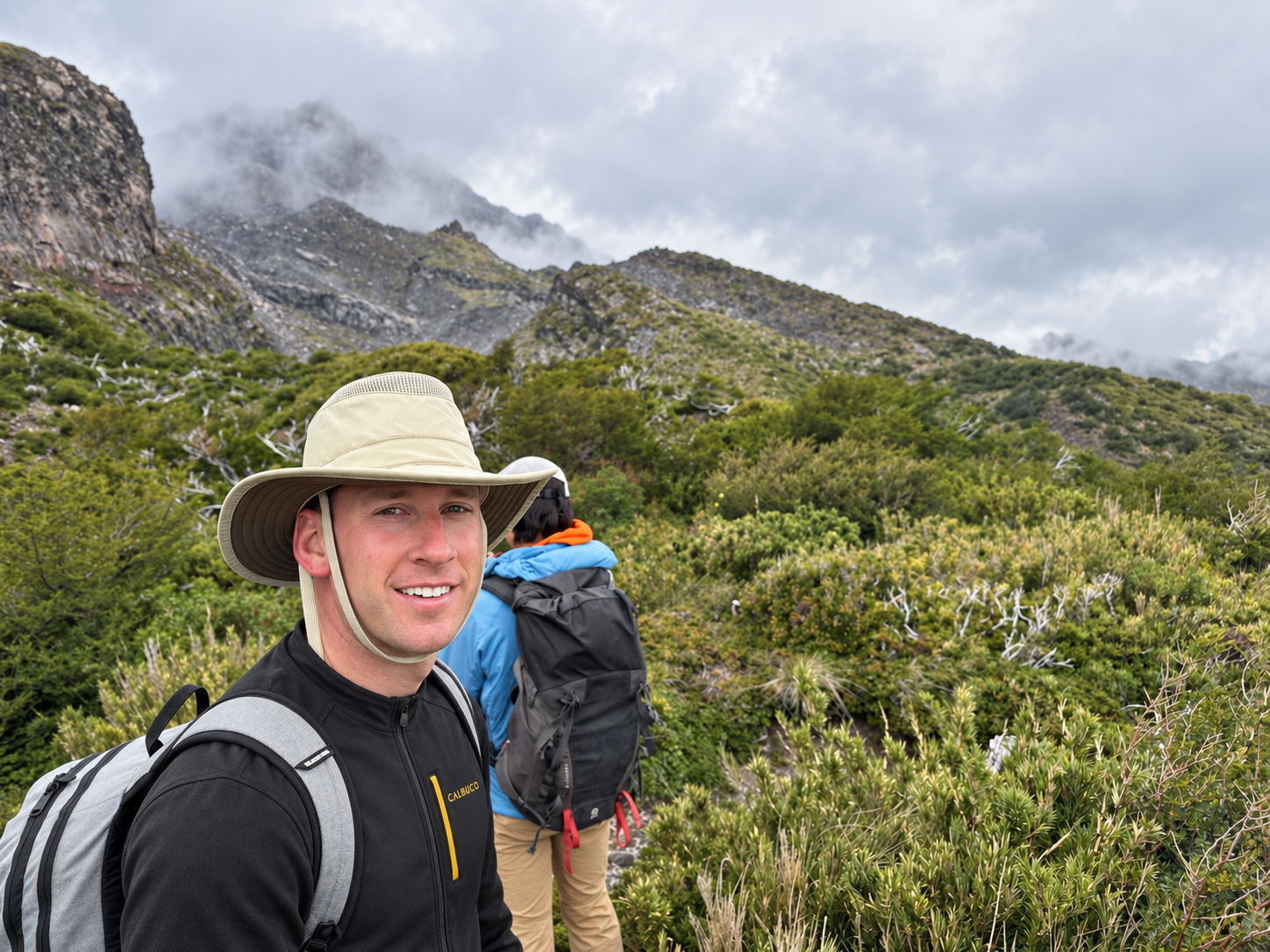 A man wearing a beige wide-brimmed hat and a black jacket with a backpack hiking in a green mountainous landscape under cloudy skies.