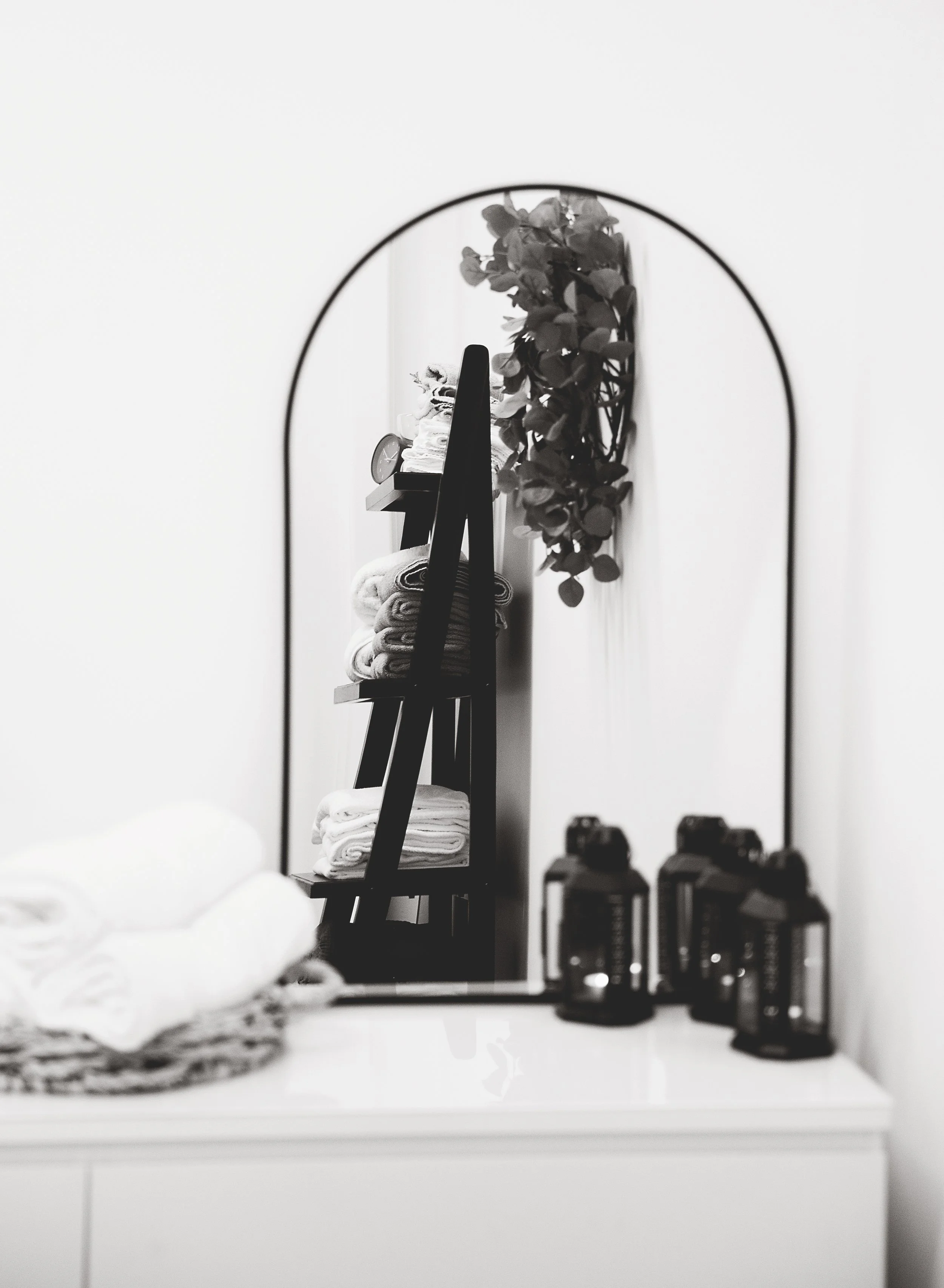 A black and white photograph of a bathroom shelf with folded towels, a clock, and a plant, viewed through an arched mirror.