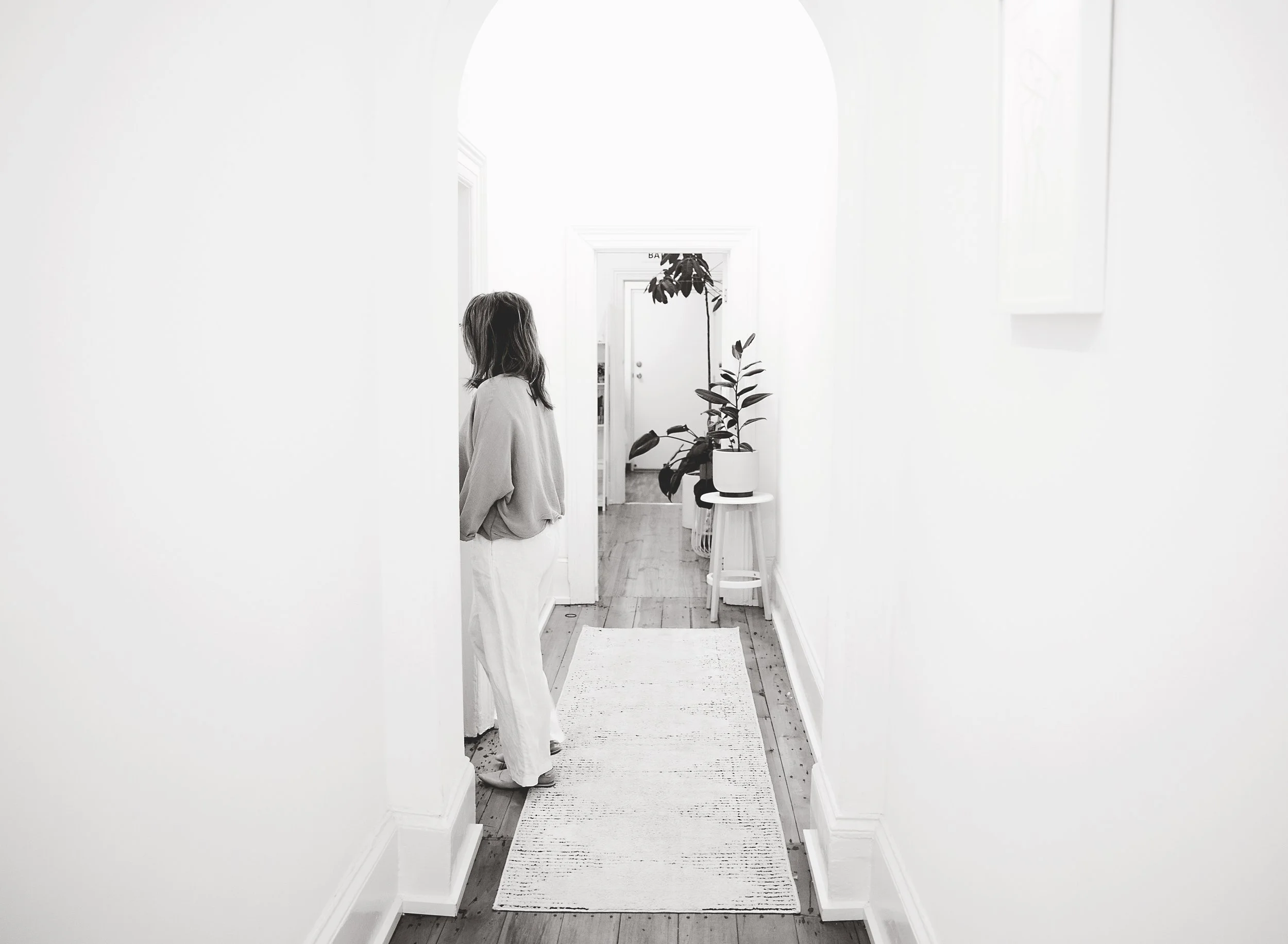 A person with long hair wearing a light-colored hoodie and pants standing in a bright, white hallway with a plant on a small table and a door at the end of the hall.