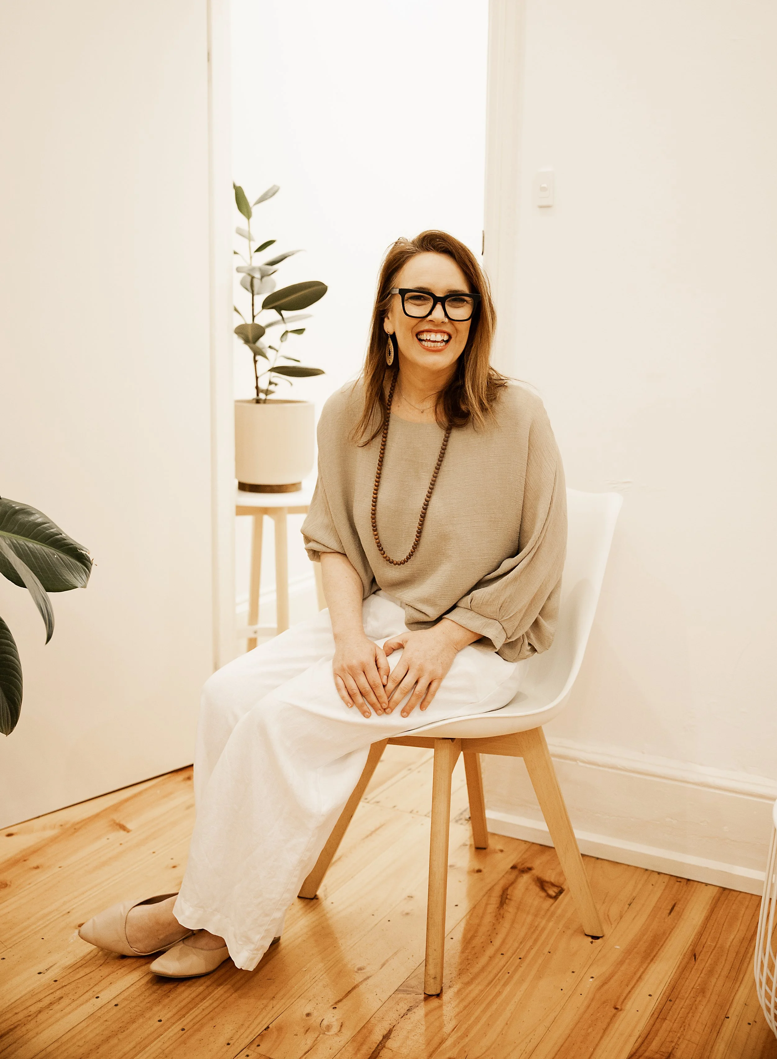 A woman with shoulder-length brown hair, wearing glasses, a beige top, white pants, and beige shoes, smiling and sitting on a wooden chair in a bright room with wooden flooring and white walls, with plants in the background.