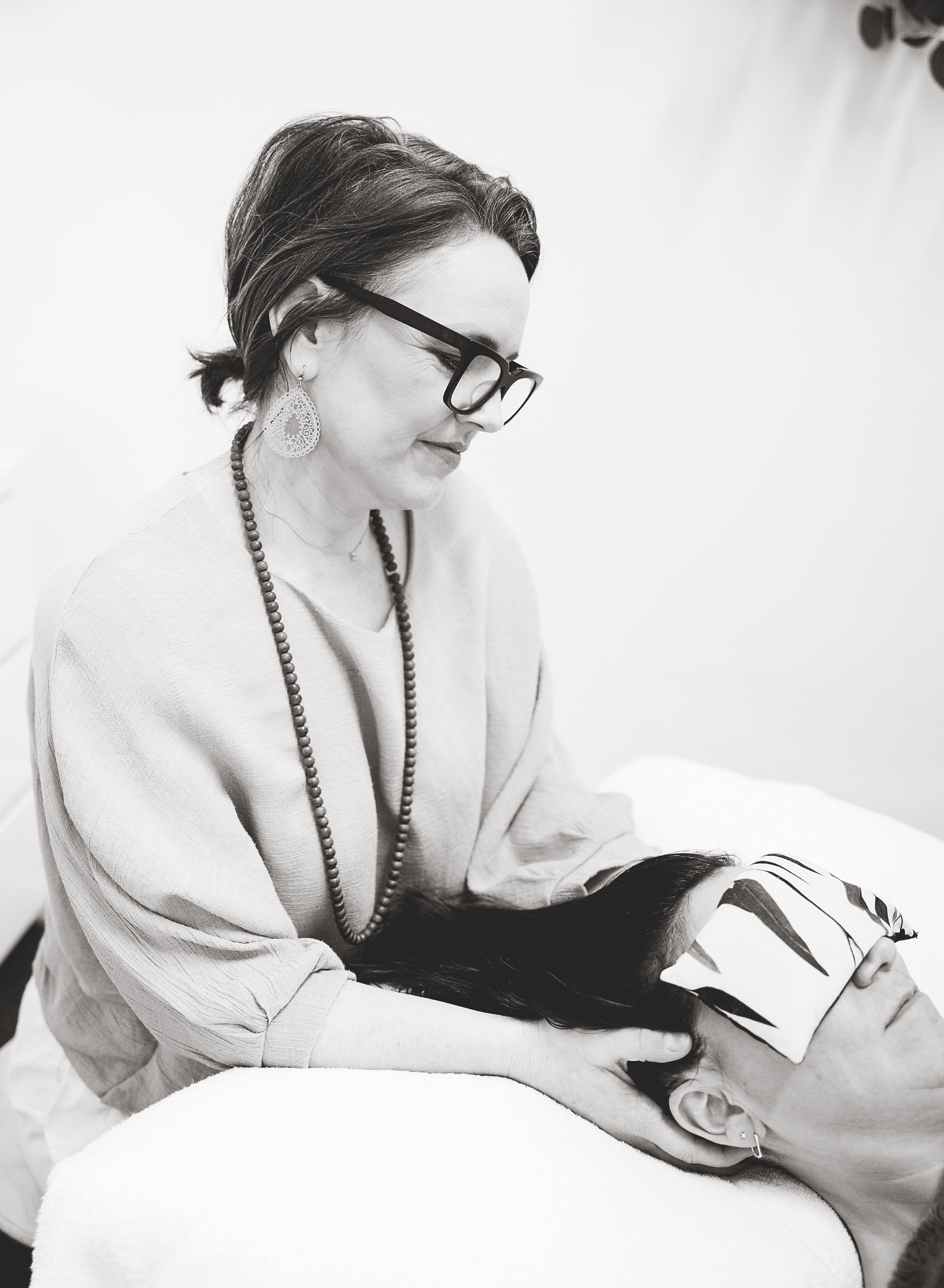 A woman with glasses and earrings appears to be receiving a head massage or treatment from another person who is lying down. The woman is smiling gently and is dressed in a light-colored top, with beaded necklaces around her neck.