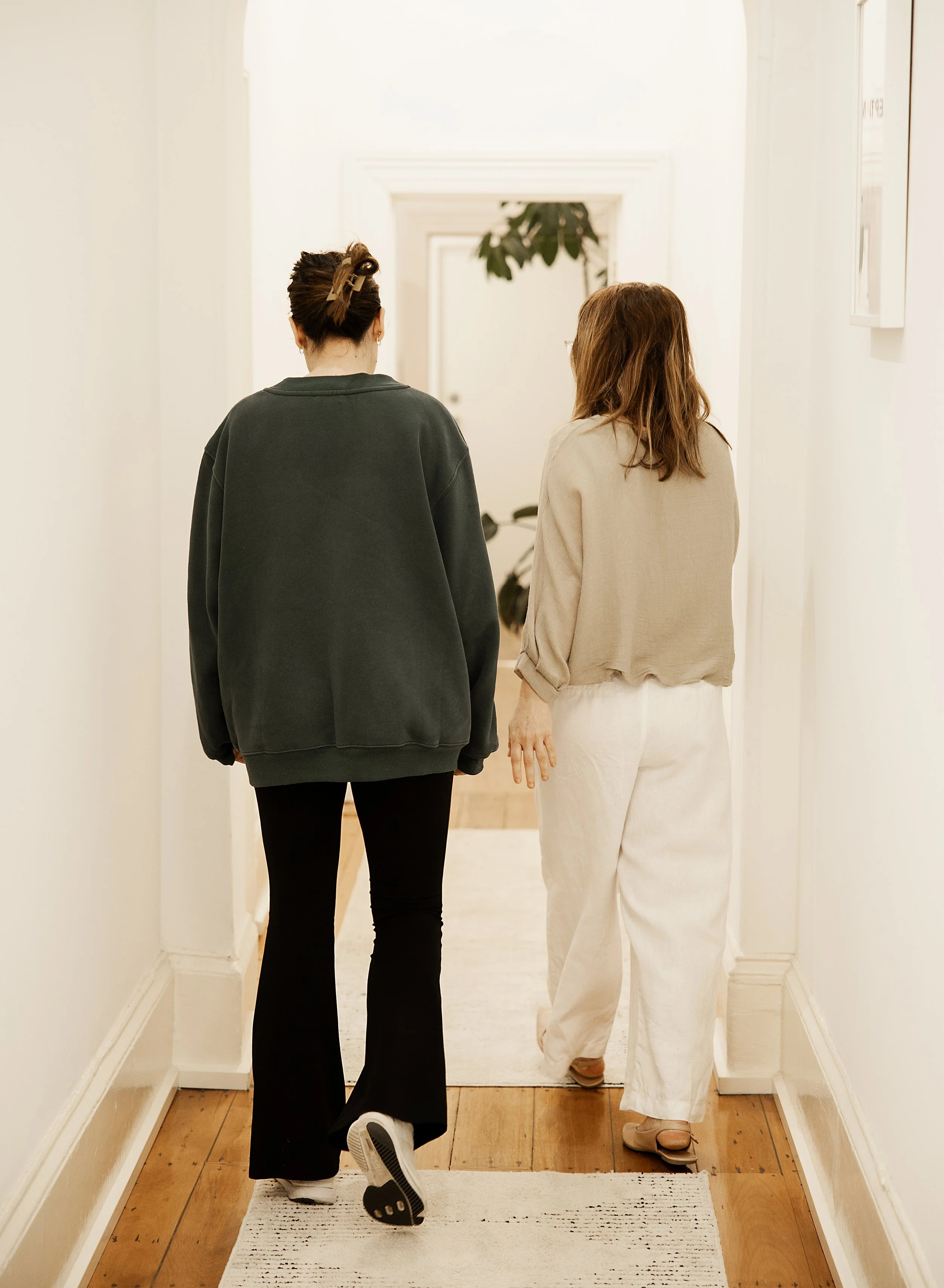 Two women walking down a hallway with beige walls and wooden flooring, one wearing a black sweatshirt and black pants, the other wearing a beige top and beige pants.