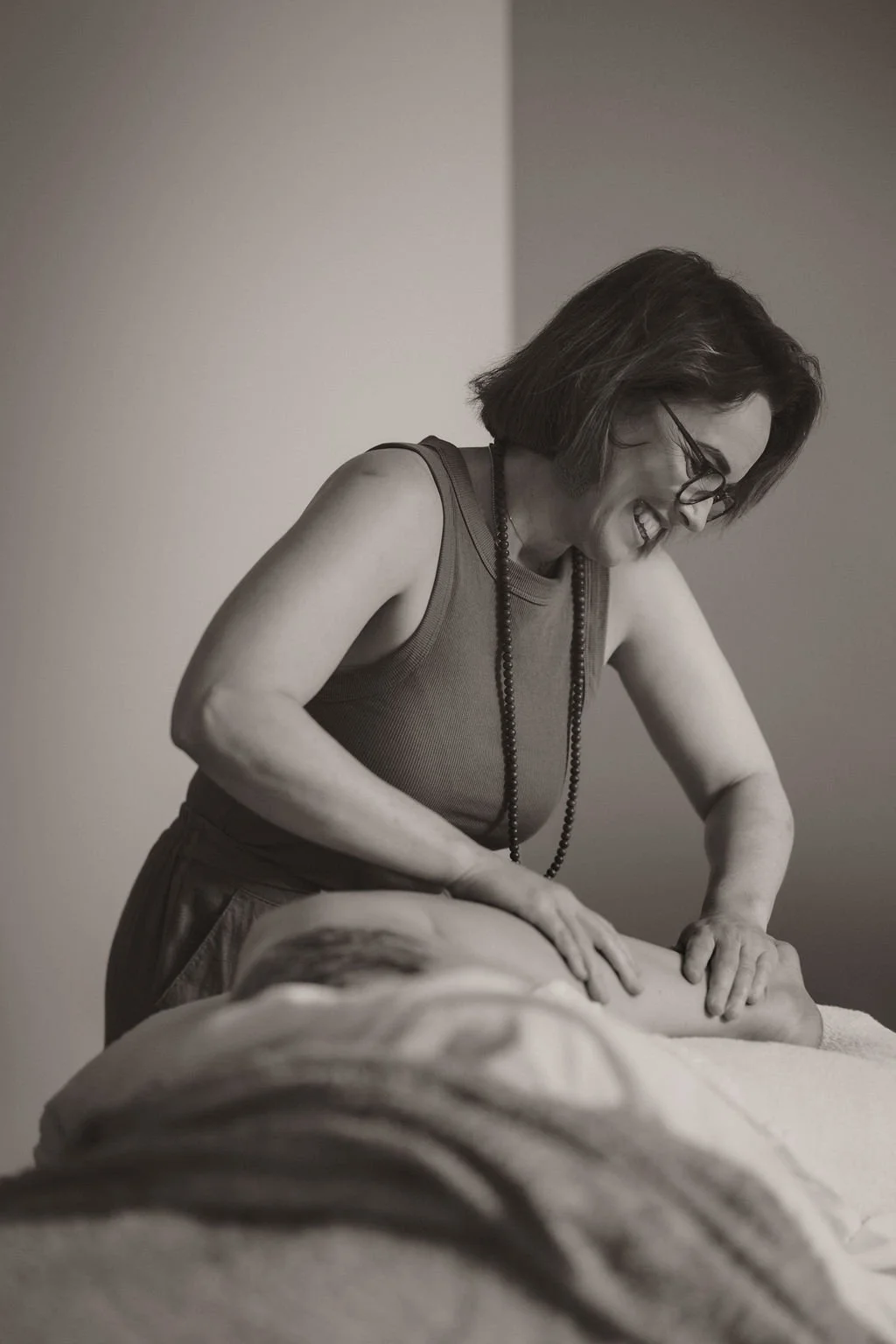 A woman with short hair, glasses, and a beaded necklace, smiling as she performs a massage on a person's leg in a therapy setting.